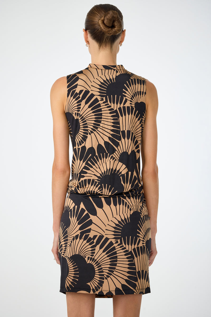 A woman with her hair in a neat bun is seen from the back wearing the Lenaya 3 Drape Dress - Juniper Jersey, a sleeveless, knee-length dress featuring a tan and black abstract fan pattern, set against a plain light background.
