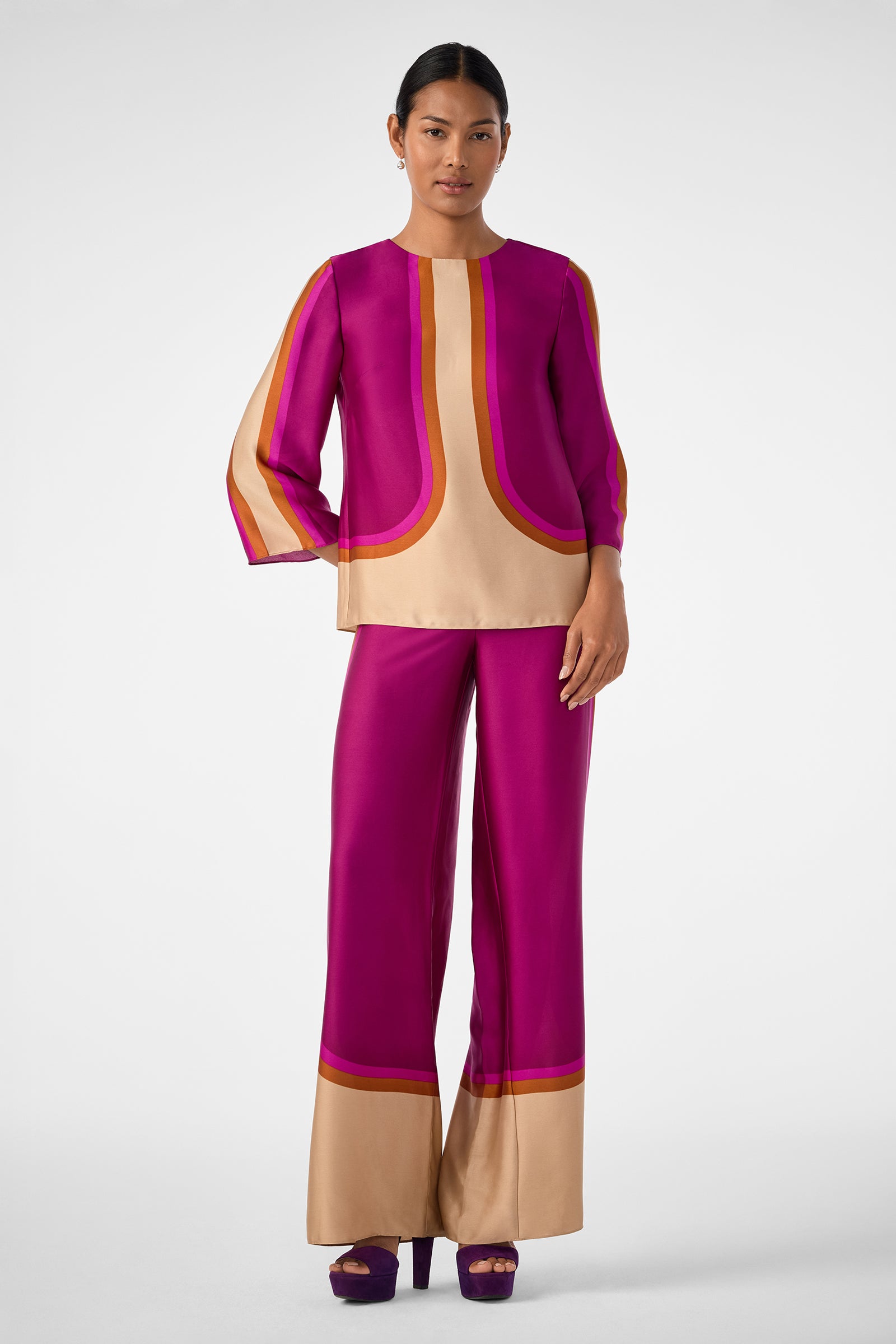 A woman stands wearing the Sprig 3 Top, a vibrant magenta and beige color-blocked long-sleeve with curved lines. She pairs it with matching wide-leg pants, purple heels, and has her hair pulled back.