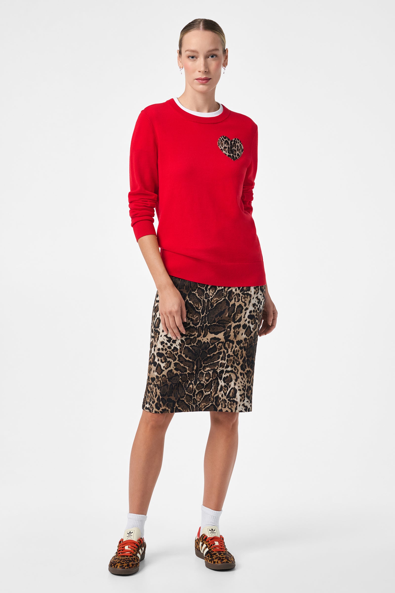 A woman stands against a white background, wearing the Jaguar Sweater with a leopard print heart, paired with a matching leopard print skirt, white socks, and leopard print sneakers with red laces. Her hair is pulled back.
