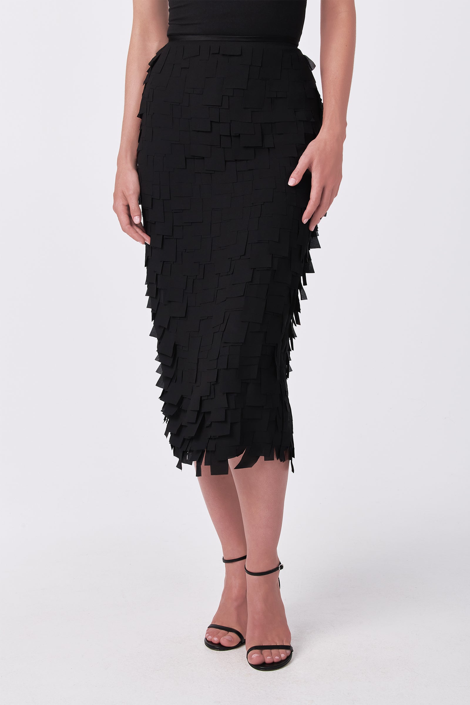 A woman wears the Dita Midi Pencil Skirt - Flutter Chiffon, featuring layered square chiffon pieces, with a black sleeveless top and strappy high-heeled sandals, standing against a white background.