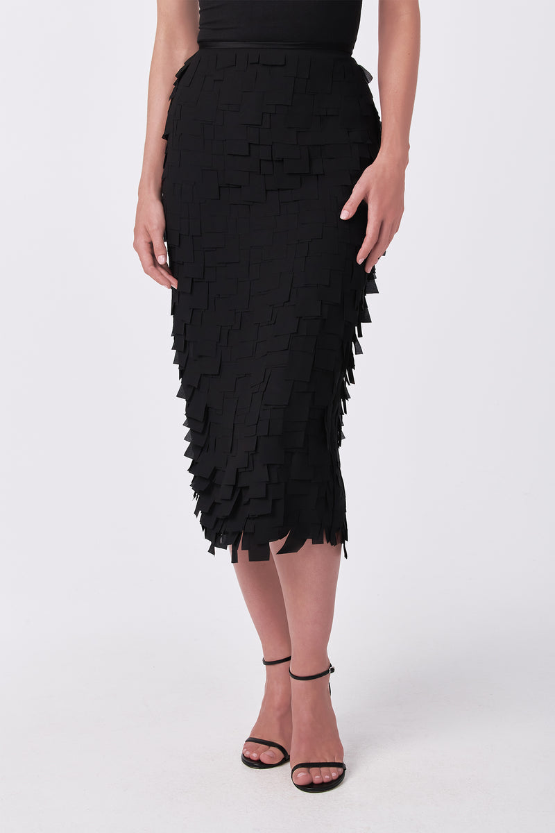 A woman wears the Dita Midi Pencil Skirt - Flutter Chiffon, featuring layered square chiffon pieces, with a black sleeveless top and strappy high-heeled sandals, standing against a white background.