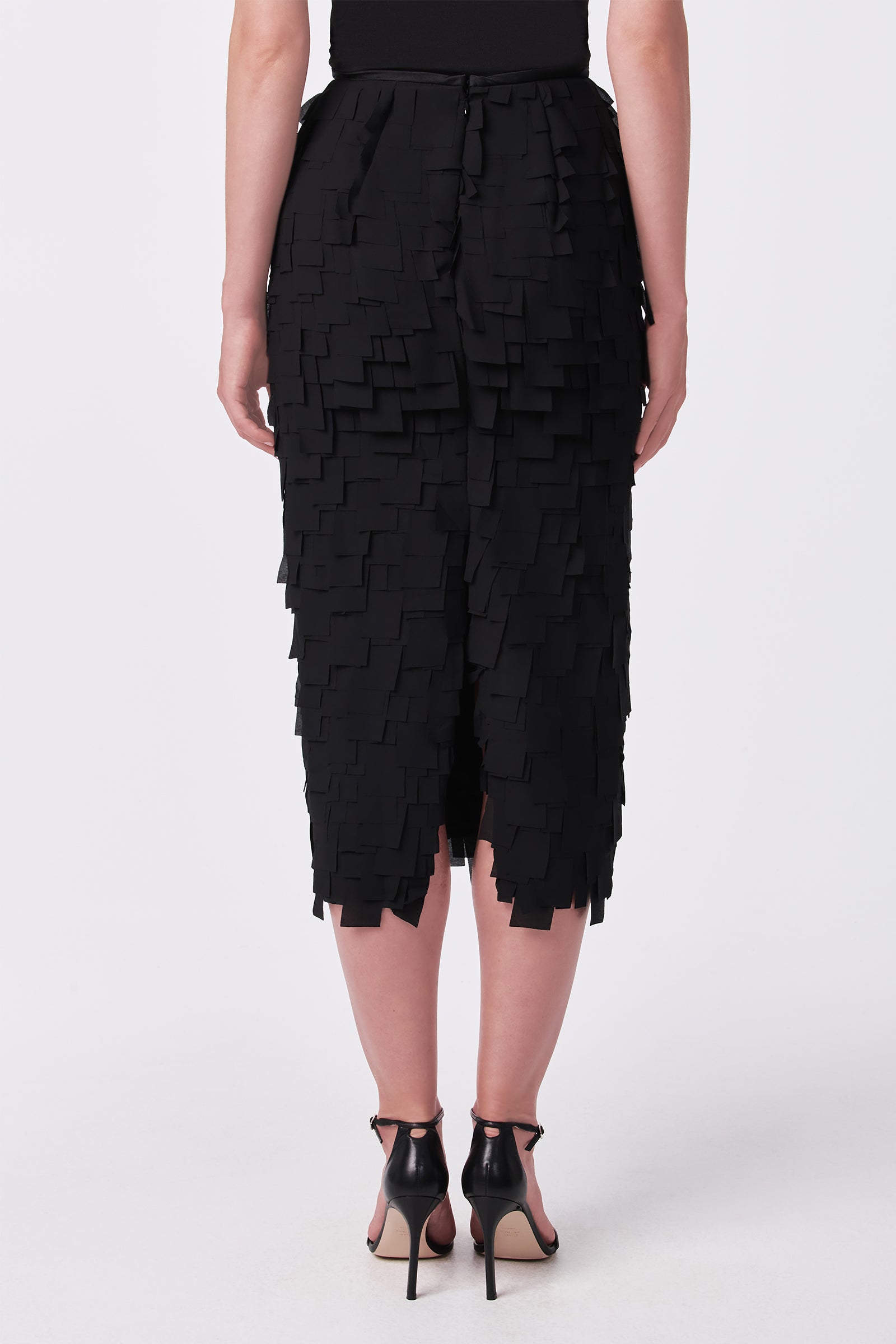 A person wearing the Dita Midi Pencil Skirt - Flutter Chiffon, featuring layered square fabric details, paired with black ankle-strap heels, photographed from the back against a white background.