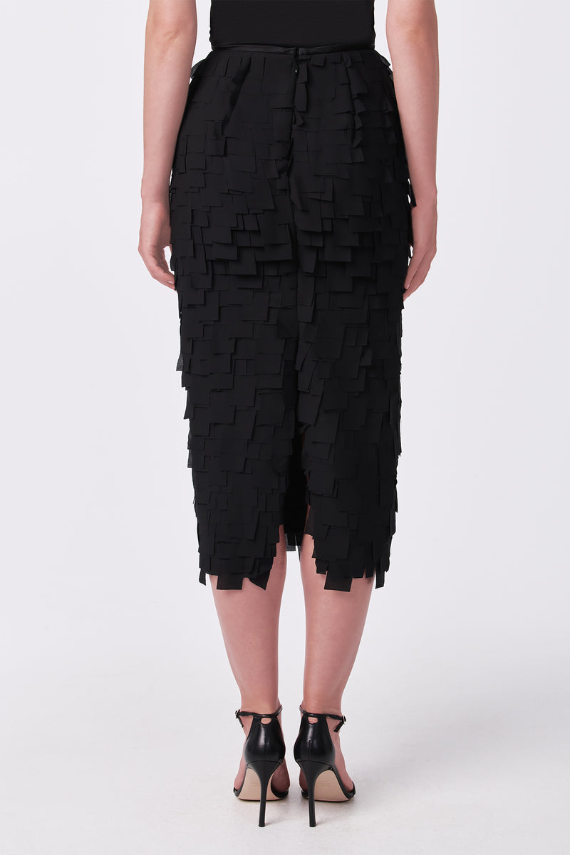 A person wearing the Dita Midi Pencil Skirt - Flutter Chiffon, featuring layered square fabric details, paired with black ankle-strap heels, photographed from the back against a white background.