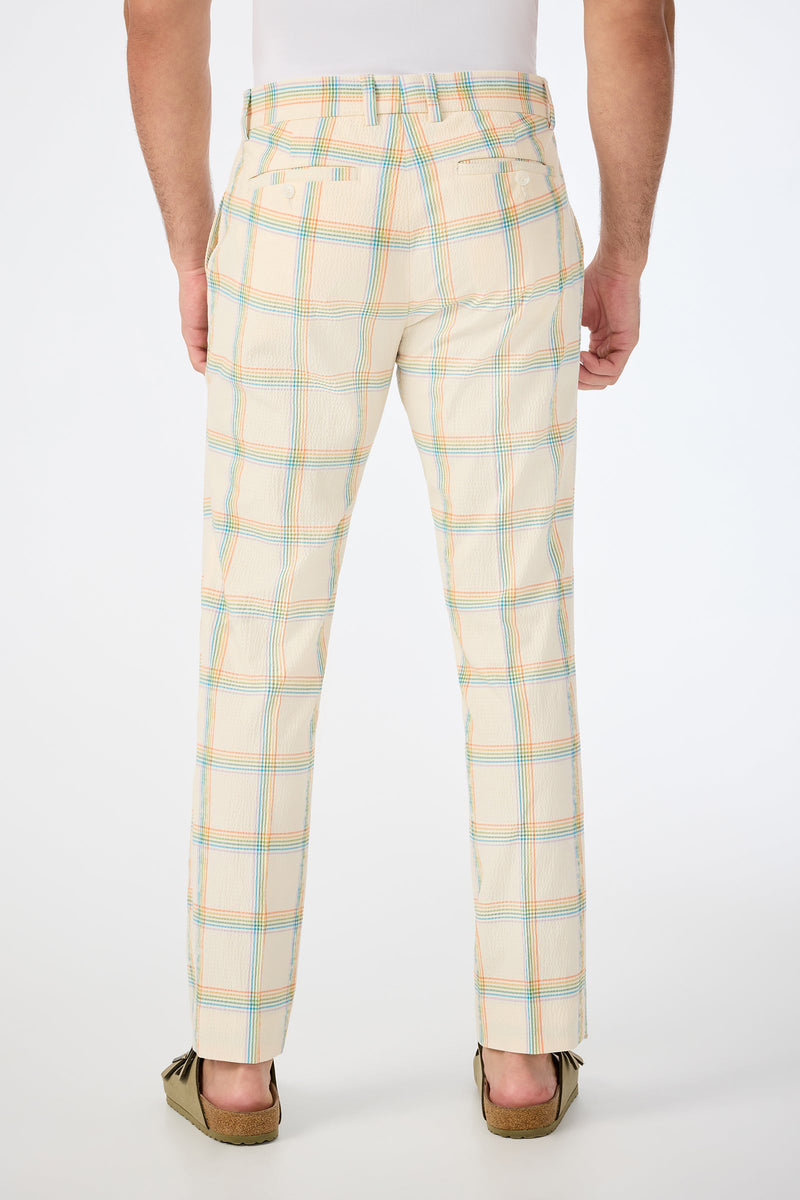A person faces away, wearing the Clyde Slim Trouser in Pride Plaid—a cream base with blue and orange plaid—paired with a tucked white shirt and brown loafers. The background is plain white.