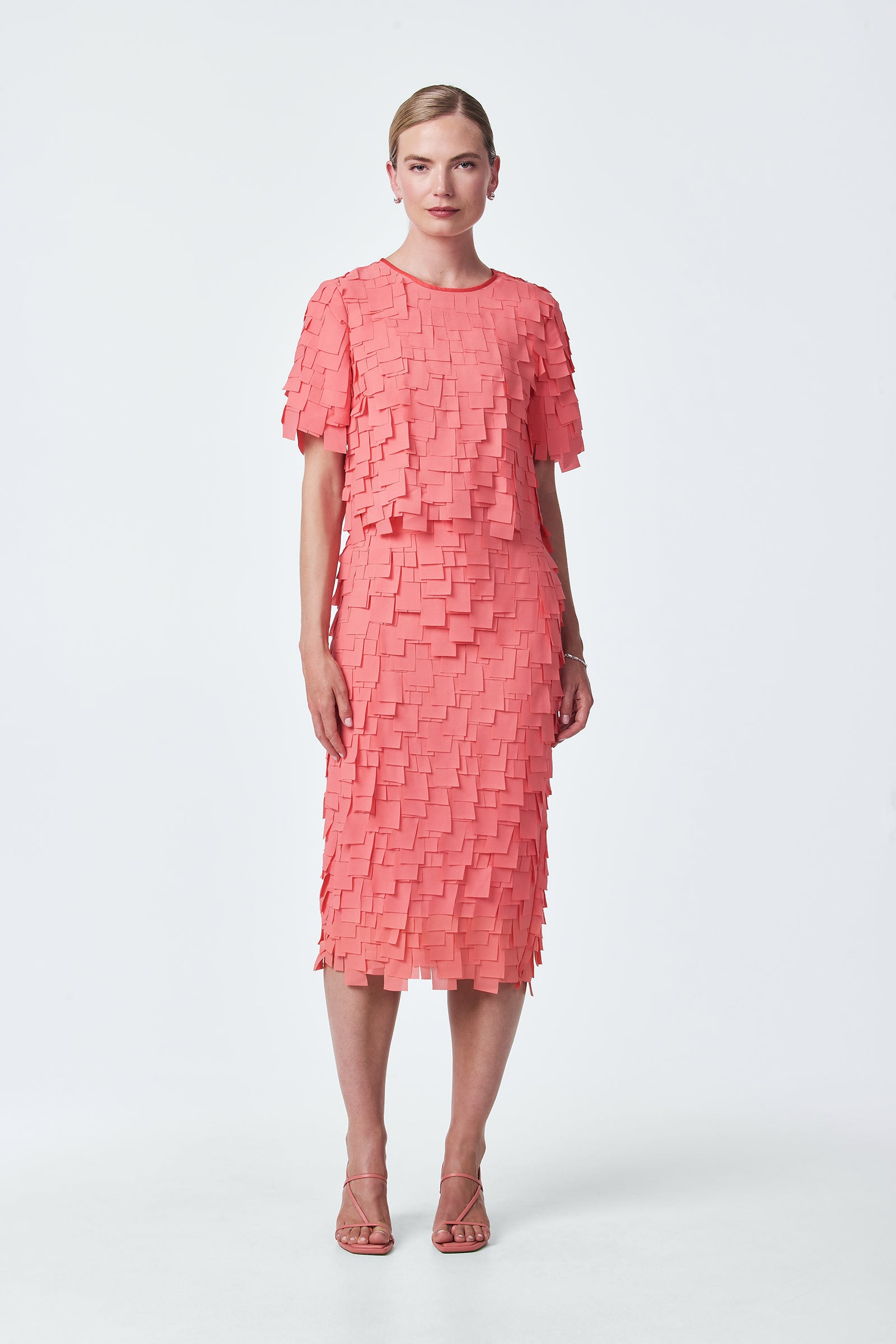 A woman with light hair pulled back and a neutral expression stands against a plain white background, wearing the Serilda Short-Sleeve Top in flutter chiffon paired with matching strappy heels.