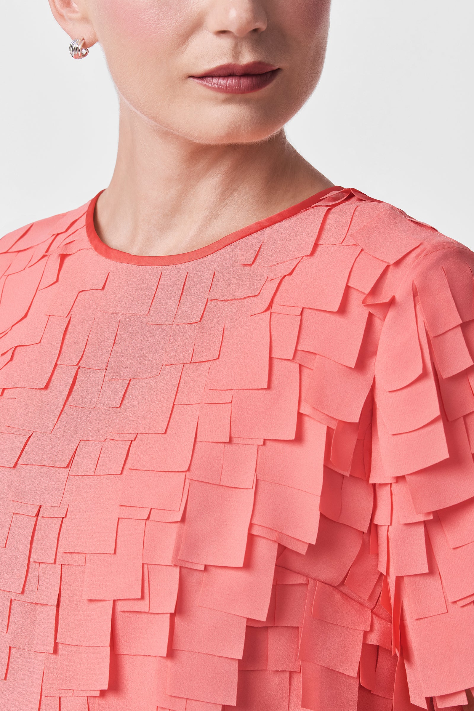 A woman wears the Serilda Short-Sleeve Top - Flutter Chiffon in coral pink, paired with light pink blush, red lipstick, and a small hoop earring. Only her lower face and upper torso are shown.