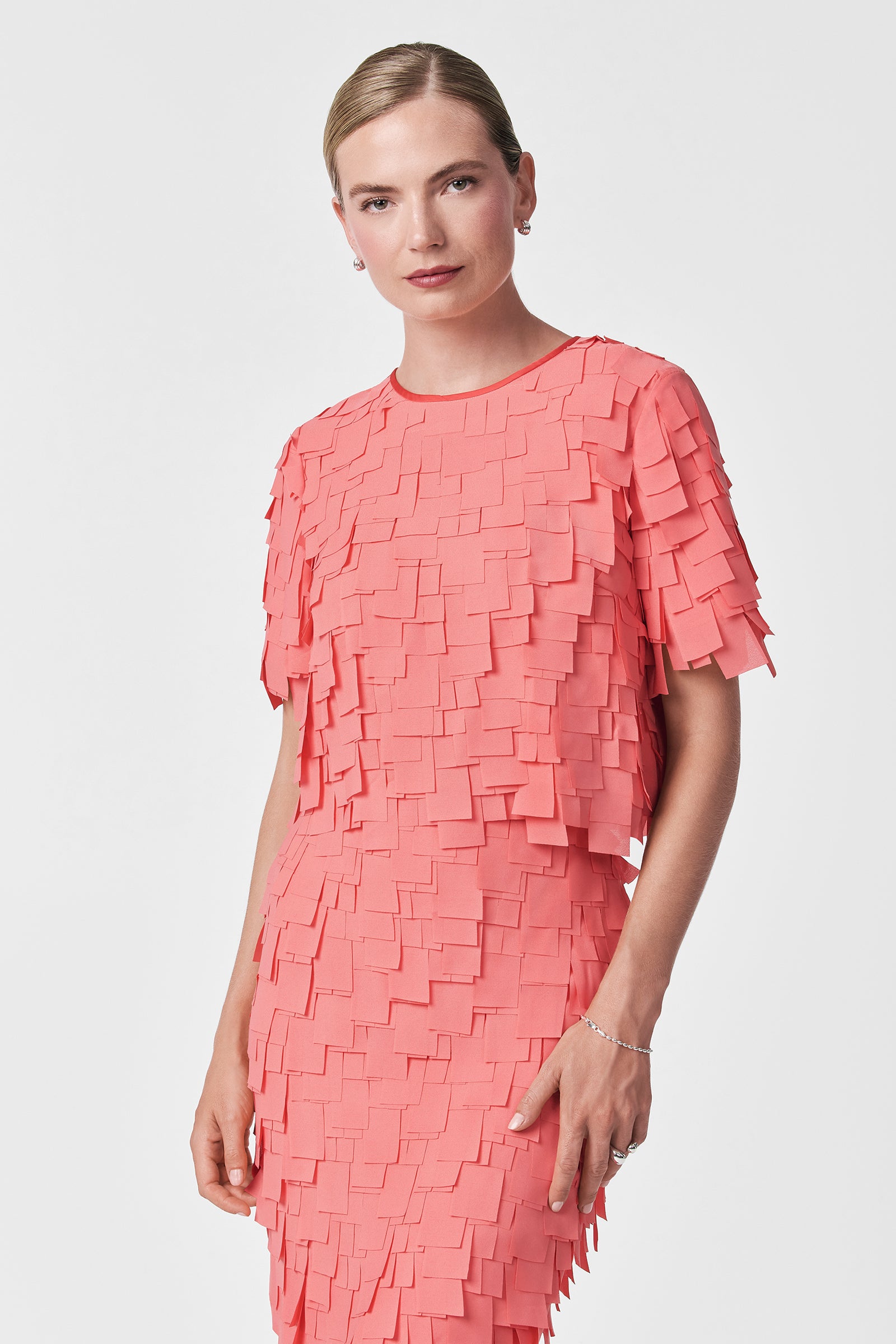 A person with light hair tied back wears the Serilda Short-Sleeve Top – Flutter Chiffon in coral pink, featuring layered, square fabric details, standing against a plain white background.