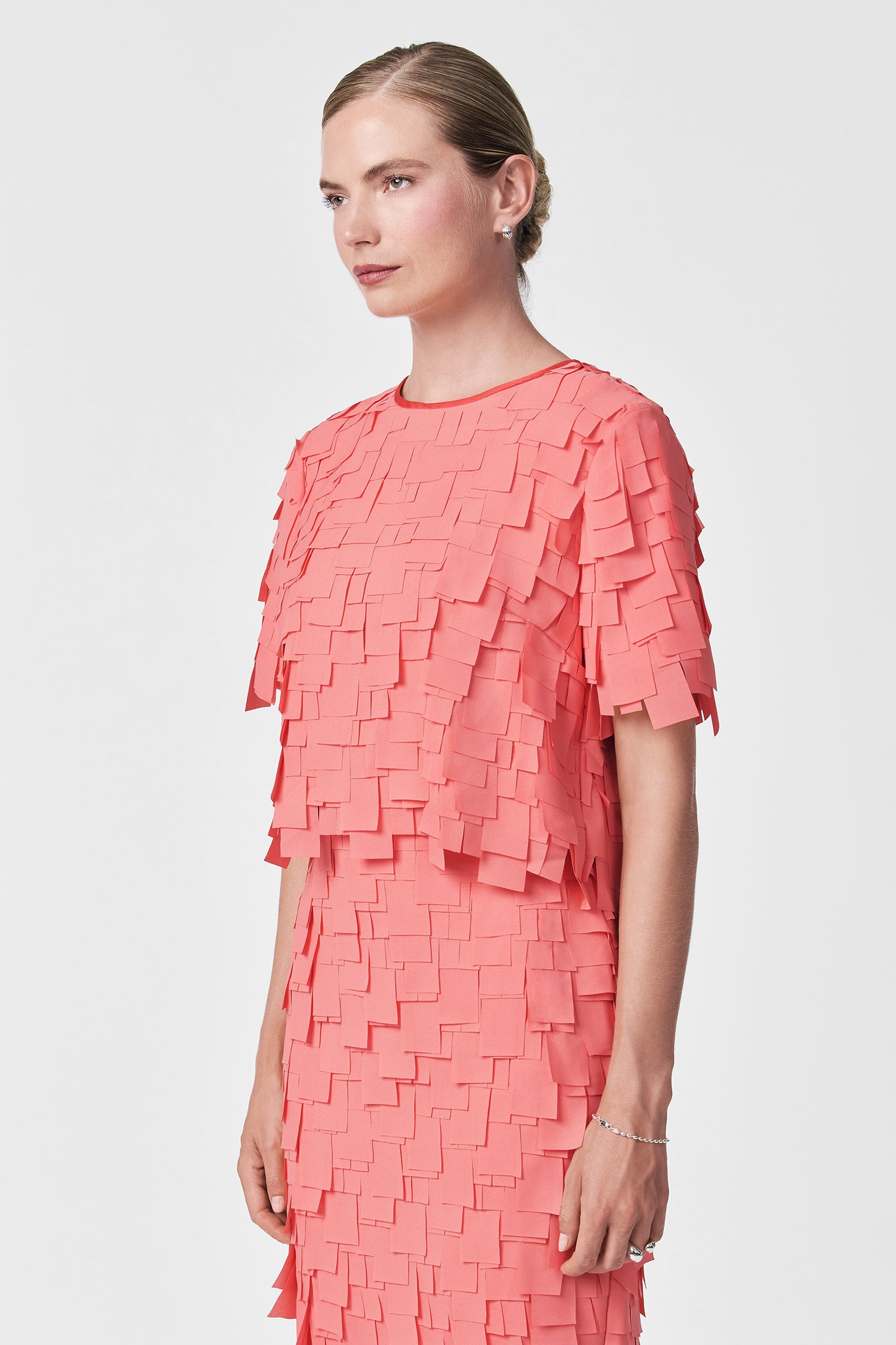 A woman with blonde hair in a low bun wears the Serilda Short-Sleeve Top – Flutter Chiffon, a coral pink style with layered, square-shaped fabric pieces, standing against a plain white background.
