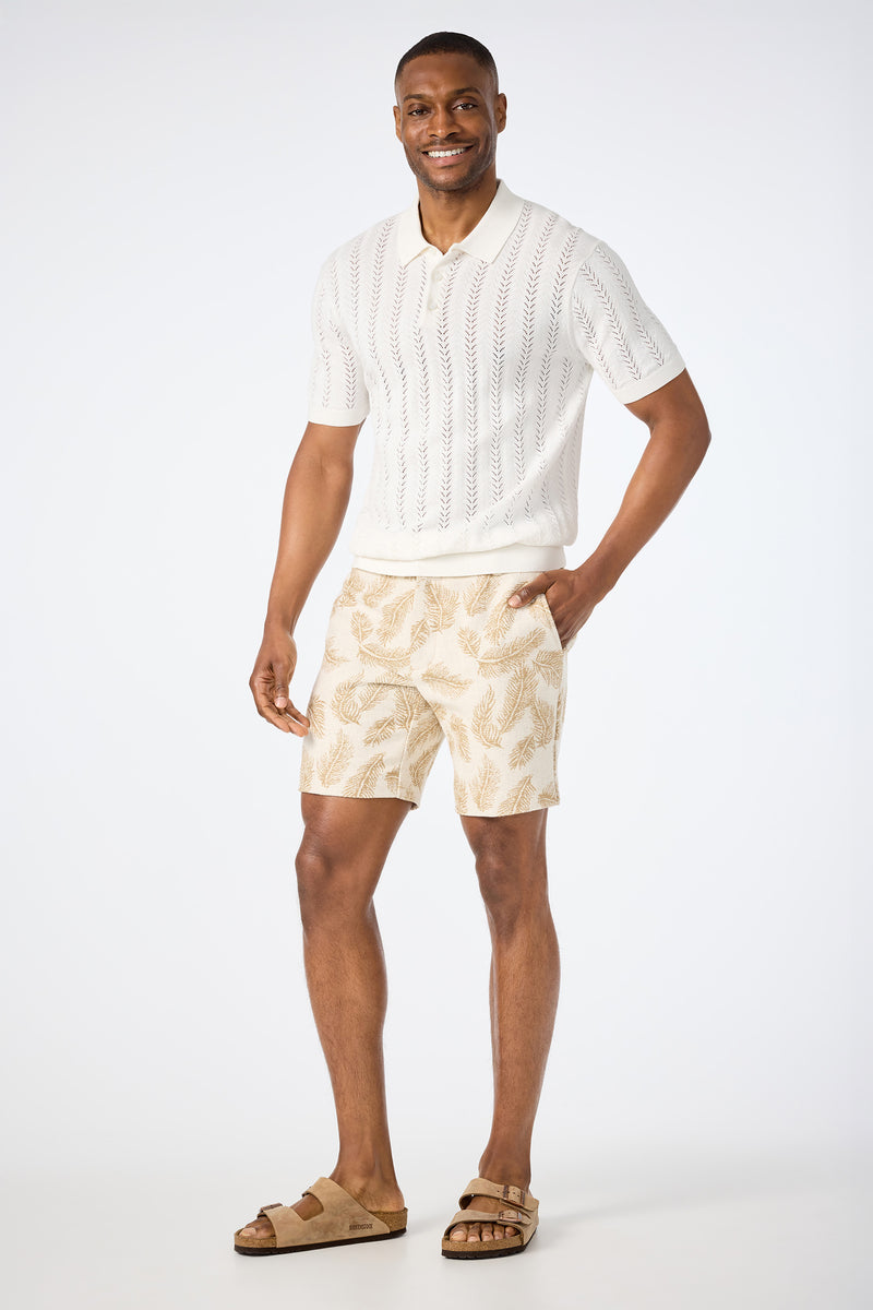 A man smiles with one hand in his pocket, wearing a white short-sleeve knit polo, tan sandals, and the Lawrence Short – Desert Fronds Jacquard against a plain white background.