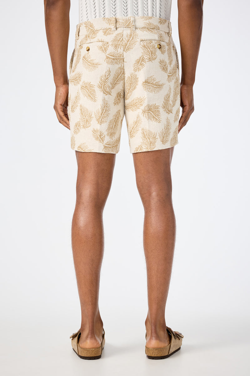 A person is seen from the back wearing the Lawrence Short - Desert Fronds Jacquard, featuring a brown leaf pattern, paired with a white textured shirt and tan slip-on shoes, standing against a plain white background.