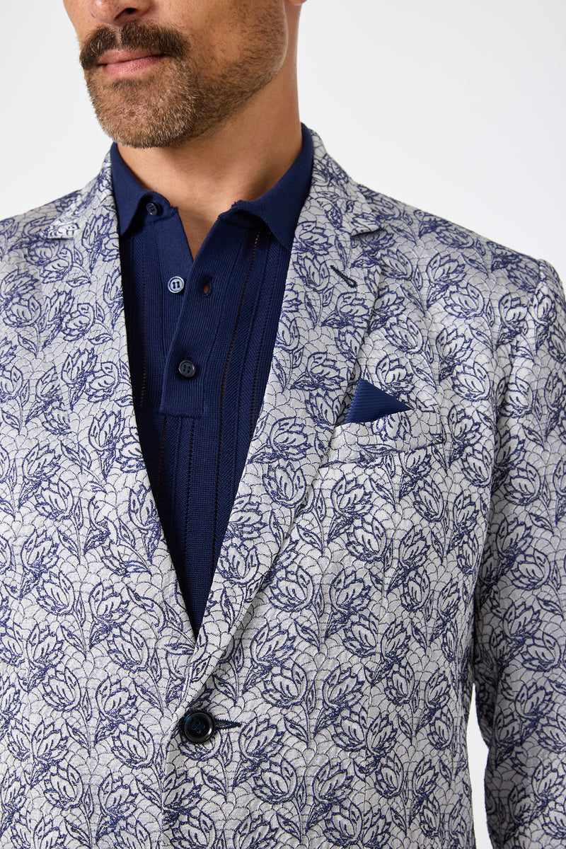 A man with a mustache is seen from his lower face to upper torso, wearing the Thurston Blazer - Efflorescent Jacquard featuring dark blue leaf patterns on light gray, paired with a navy collared shirt and matching pocket square.