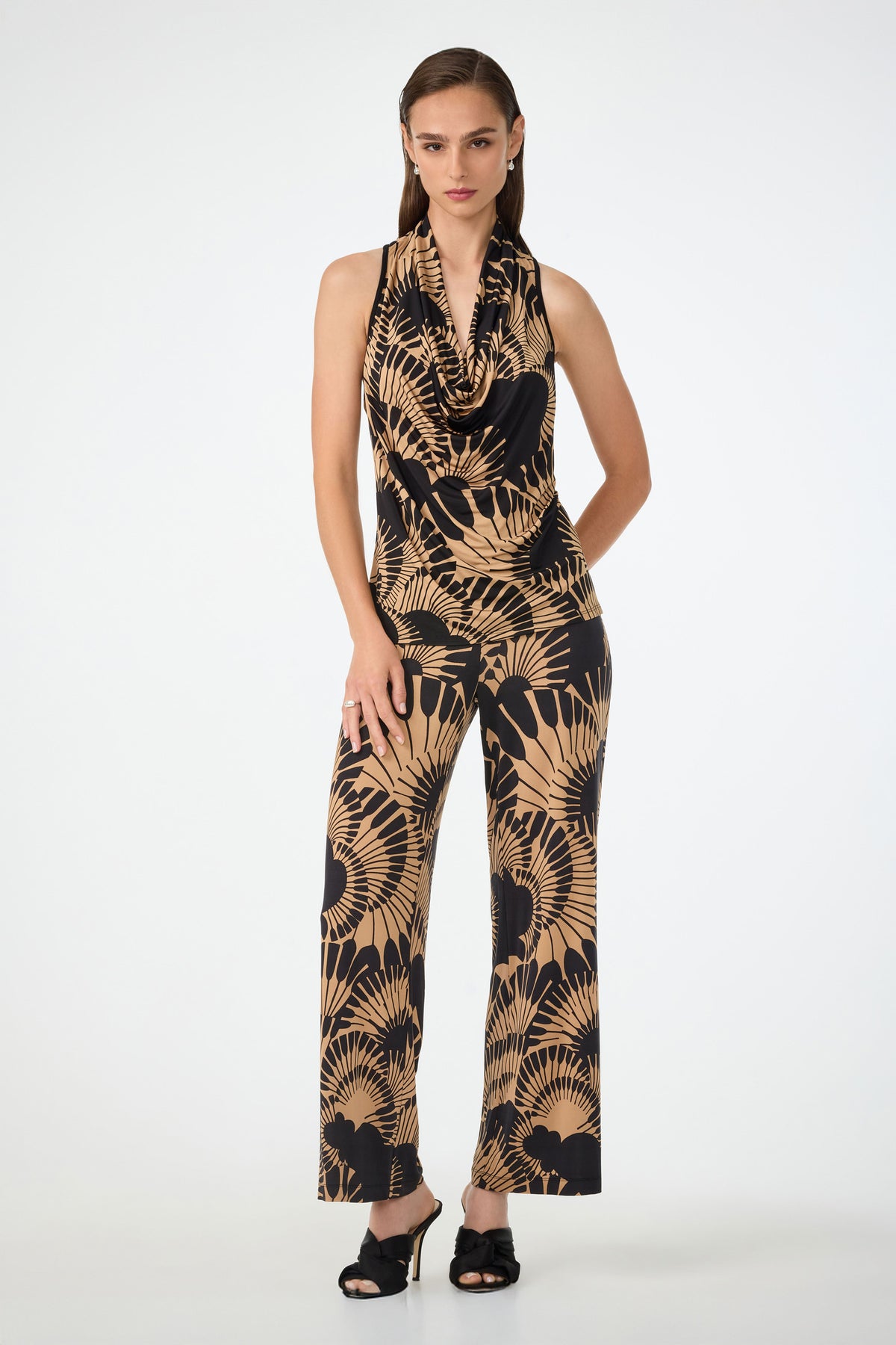 A woman with straight brown hair stands against a plain background wearing the Mosiac Cowl Neck Top in Juniper Jersey, featuring a sleeveless design and bold black and tan leaf pattern, paired with black open-toe heels.