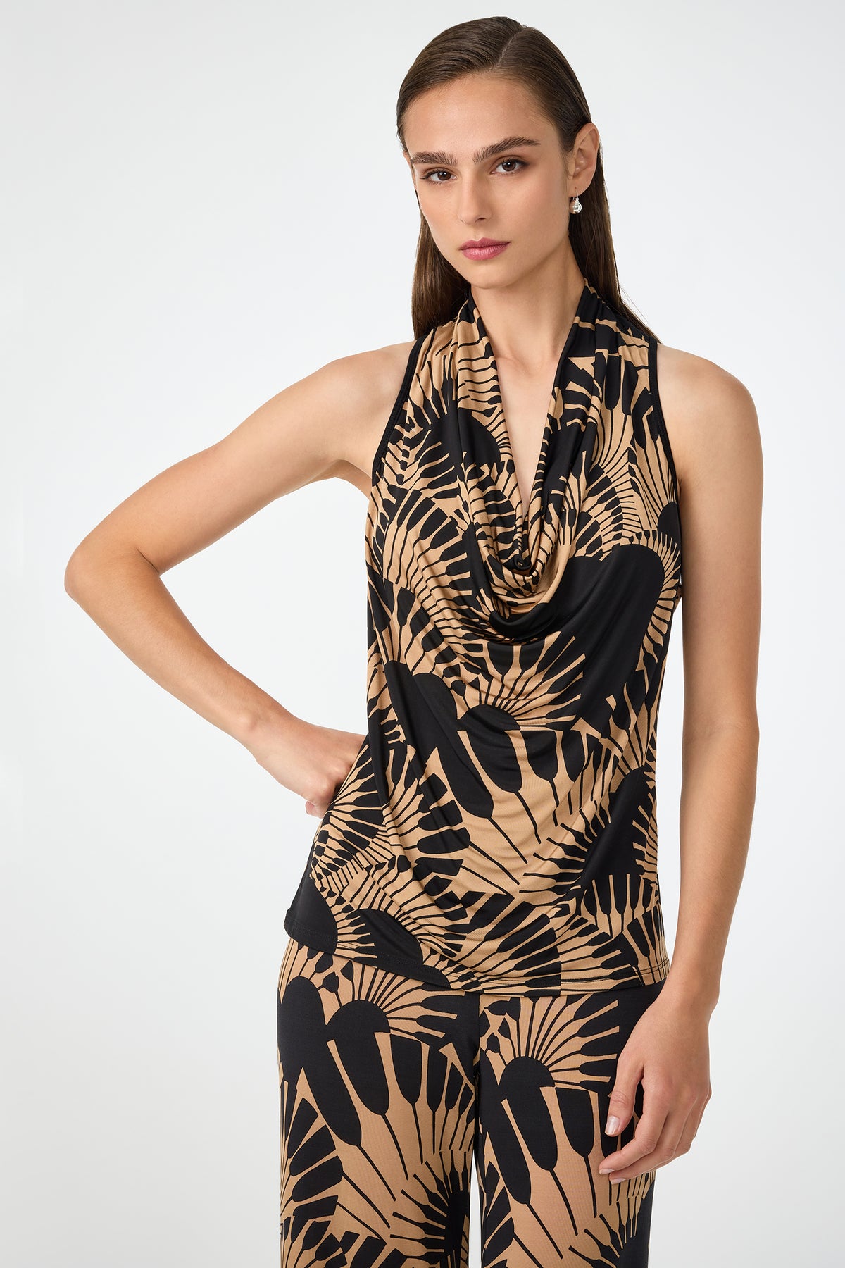 A woman with straight brown hair wears the Mosiac Cowl Neck Top in Juniper Jersey, paired with matching pants featuring a bold geometric pattern. She poses with one hand on her hip and a neutral expression against a plain background.