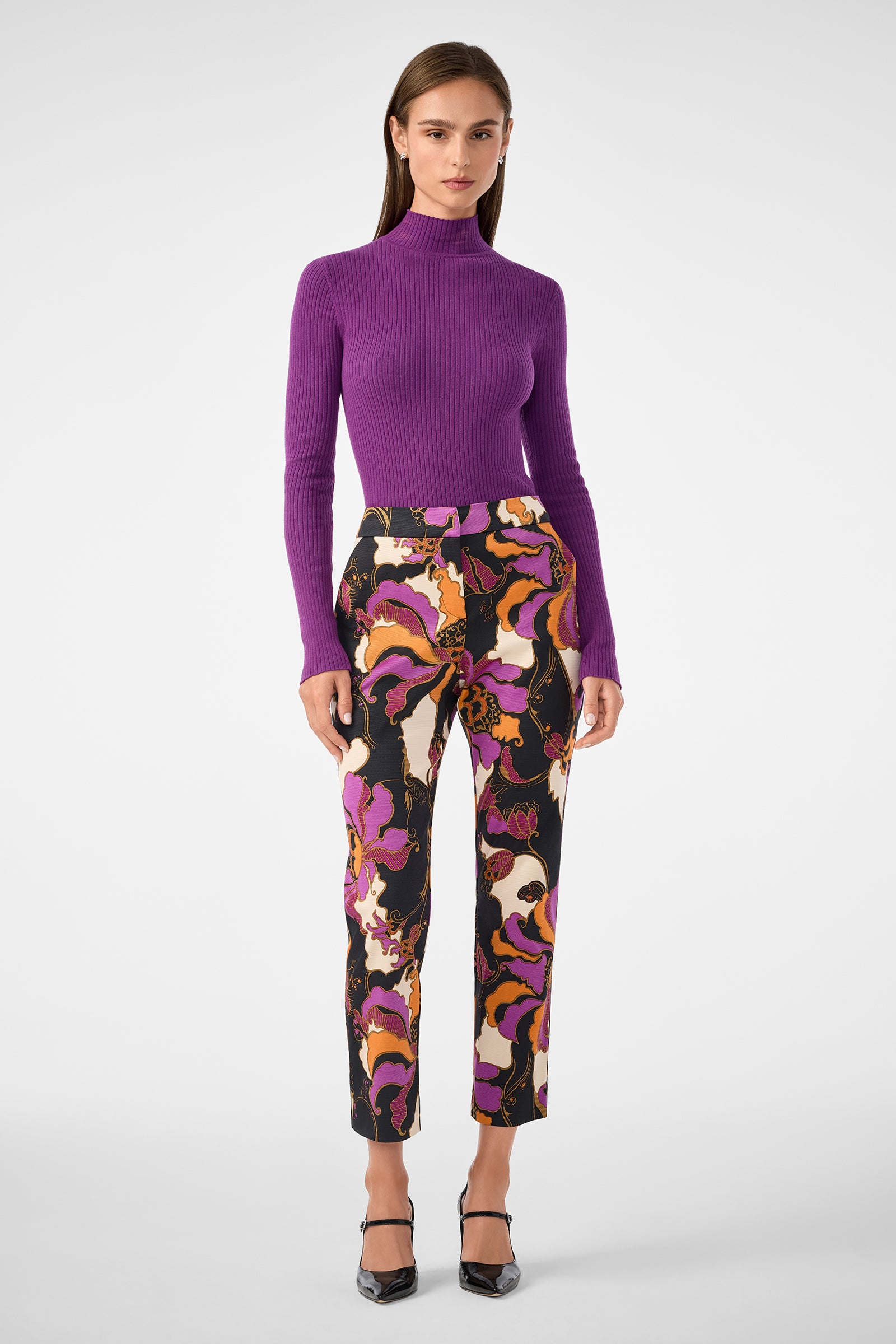 A woman stands facing forward, wearing a fitted purple ribbed turtleneck sweater, black pointed-toe heels, and the Moss 2 Pant—high-waisted pants featuring colorful abstract prints in black, orange, pink, and white.