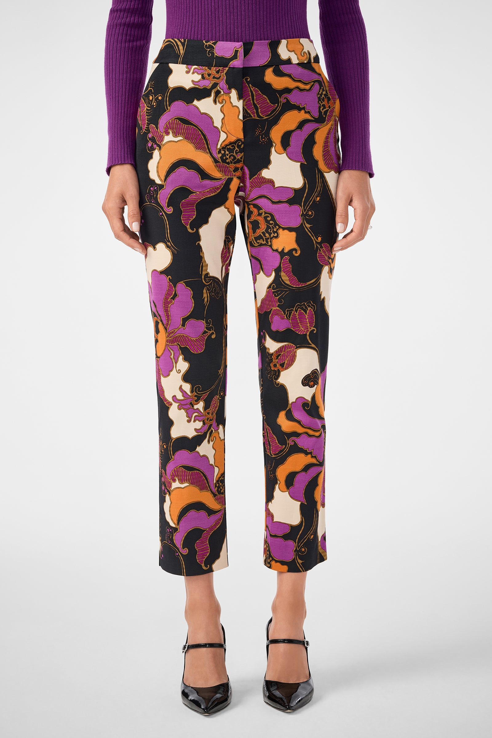 Someone wearing the Moss 2 Pant—high-waisted, straight-leg pants with a bold purple, pink, orange, and black floral pattern—paired with a purple ribbed sweater and black pointed-toe heels.