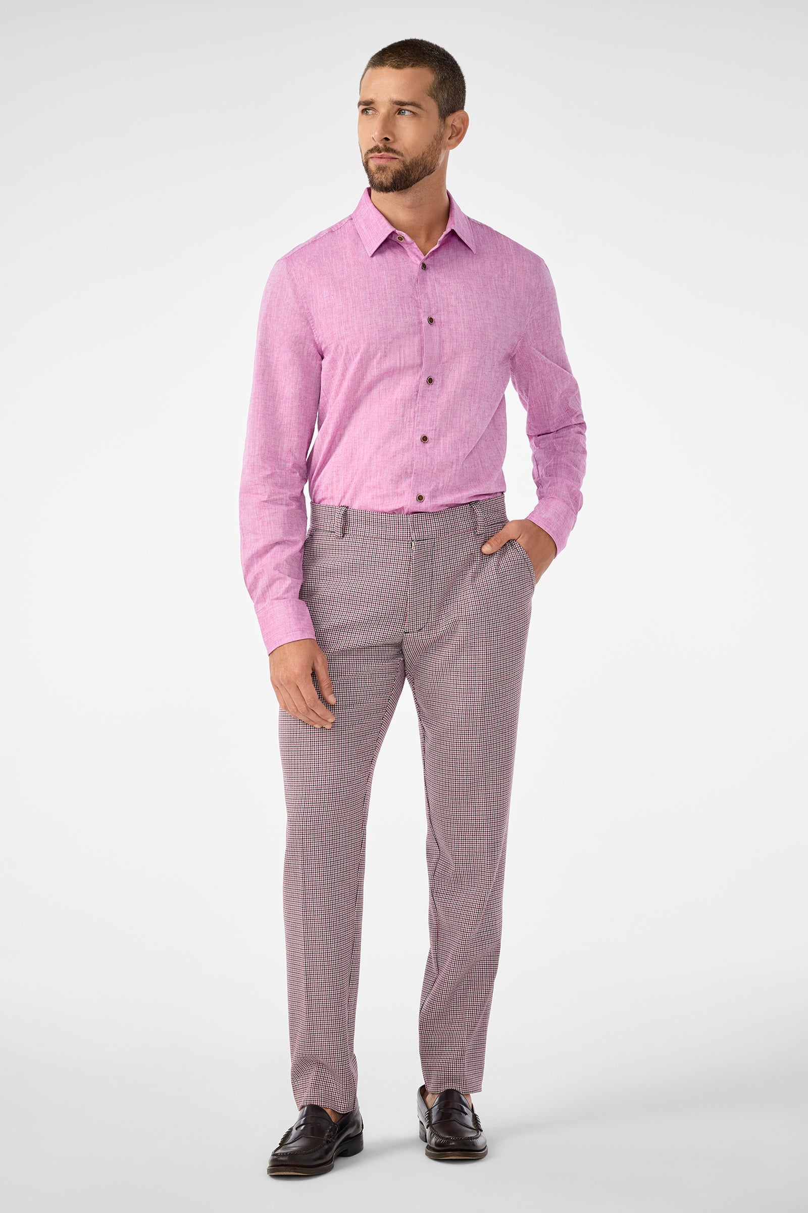 A bearded man stands slightly left, wearing the Aaron Slim Fit Long-Sleeve Shirt in Geranium tucked into patterned dress pants with one hand in his pocket and dark loafers, against a plain white background.