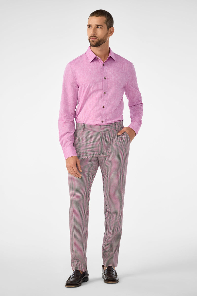 A bearded man stands slightly left, wearing the Aaron Slim Fit Long-Sleeve Shirt in Geranium tucked into patterned dress pants with one hand in his pocket and dark loafers, against a plain white background.