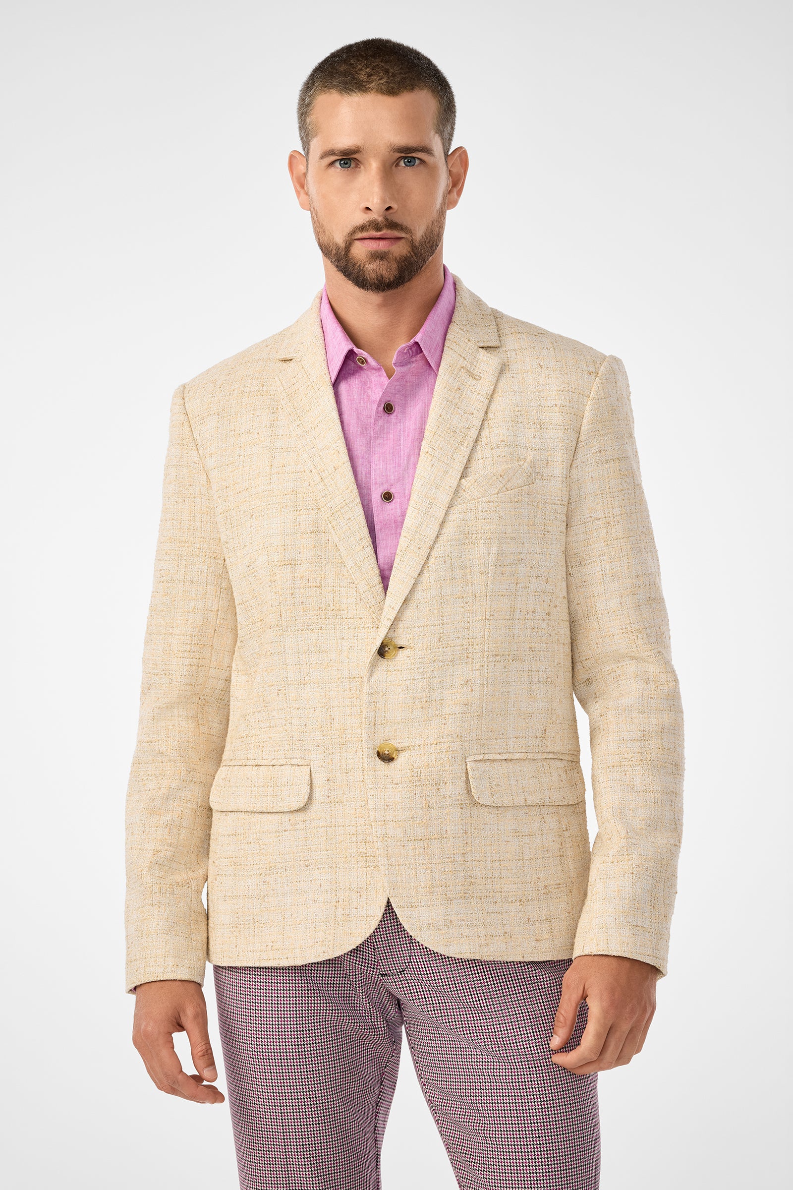 A man with short hair and a beard wears the Thurston Blazer, a light beige textured piece, over a pink button-up shirt and patterned trousers, standing against a plain white background.