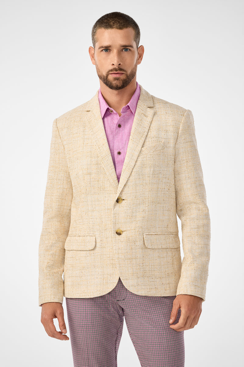A man with short hair and a beard wears the Thurston Blazer, a light beige textured piece, over a pink button-up shirt and patterned trousers, standing against a plain white background.