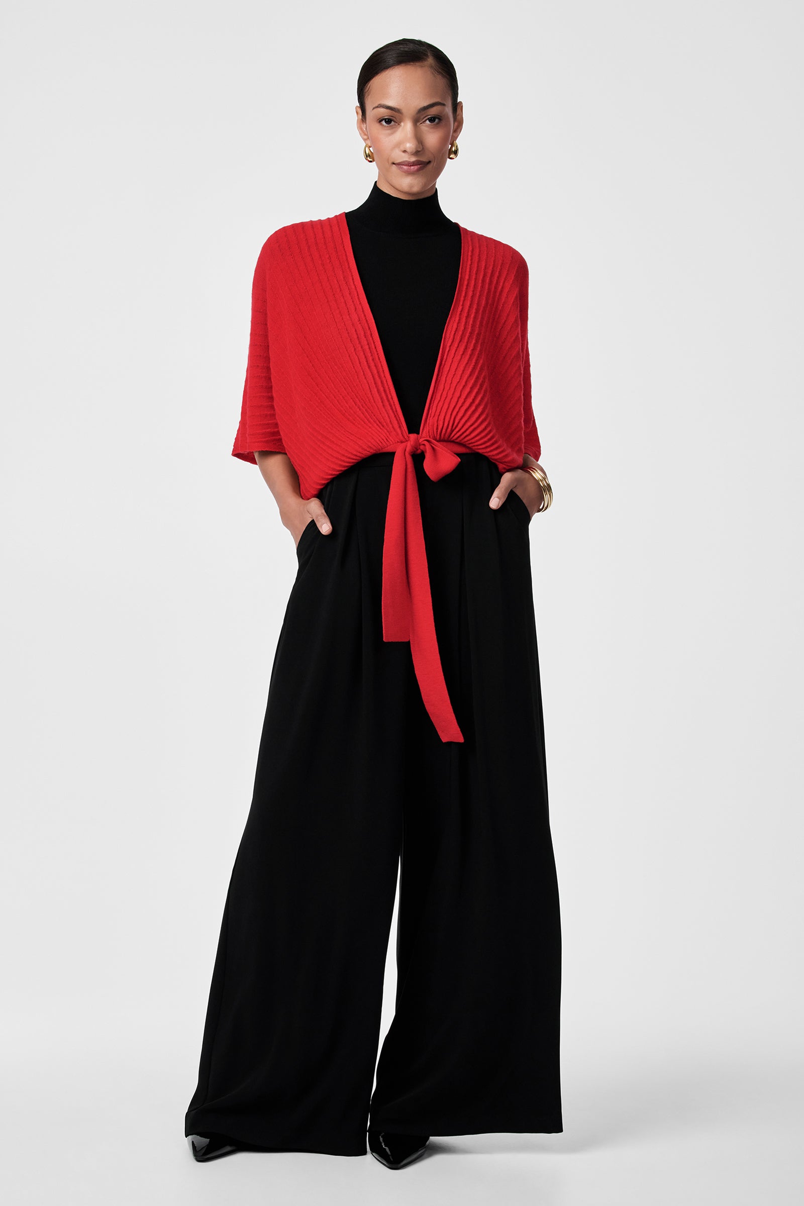 A woman with short hair wears the Exceptional Tie Front Sweater in red over a black turtleneck and wide-leg pants, accessorized with small earrings and a gold bracelet, posing against a plain white background.