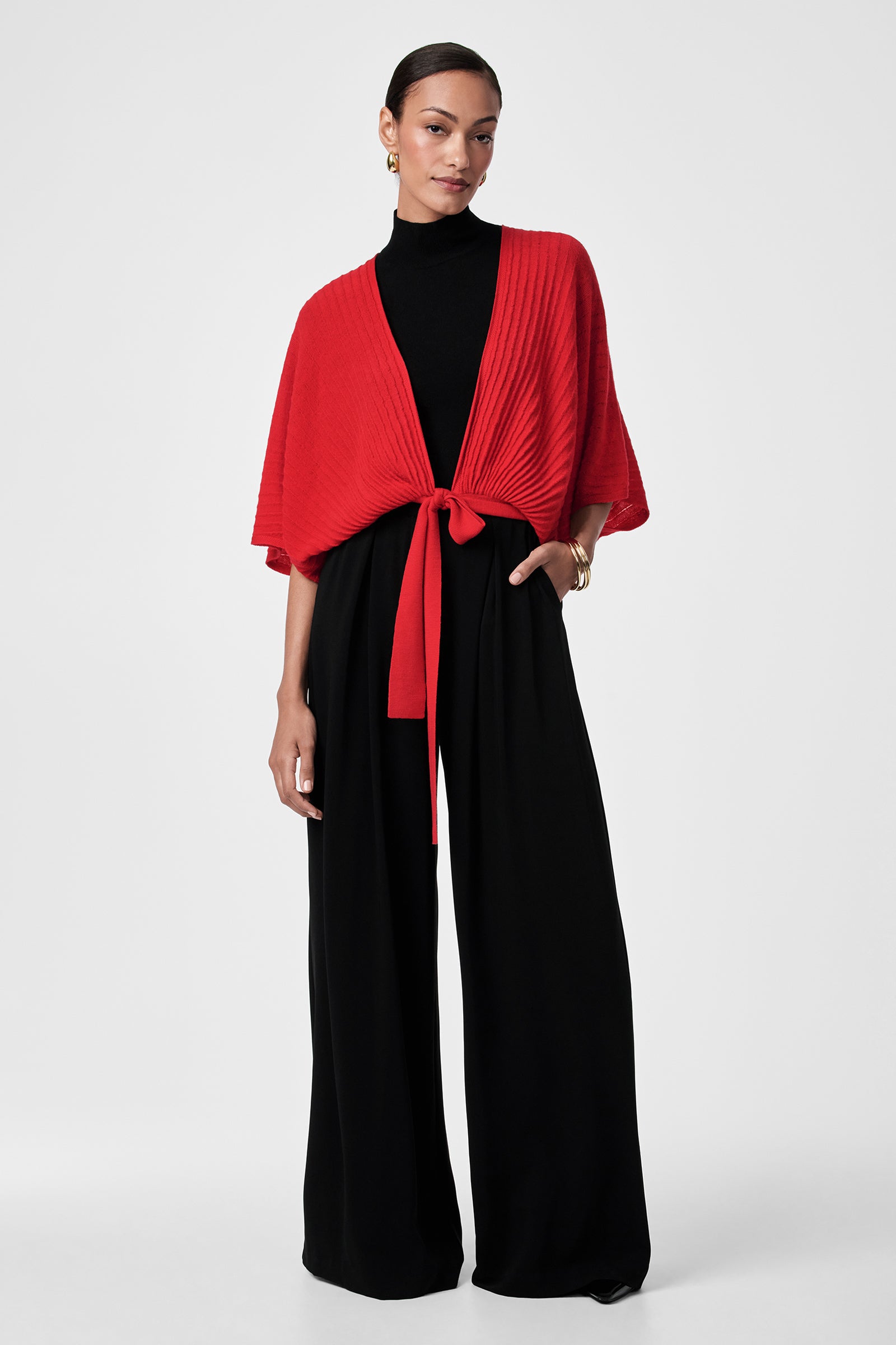 A woman stands against a plain background, wearing a black wide-leg jumpsuit and the Exceptional Tie Front Sweater in red, tied at the waist. She accessorizes with gold hoop earrings, a gold bracelet, and her hair is pulled back.