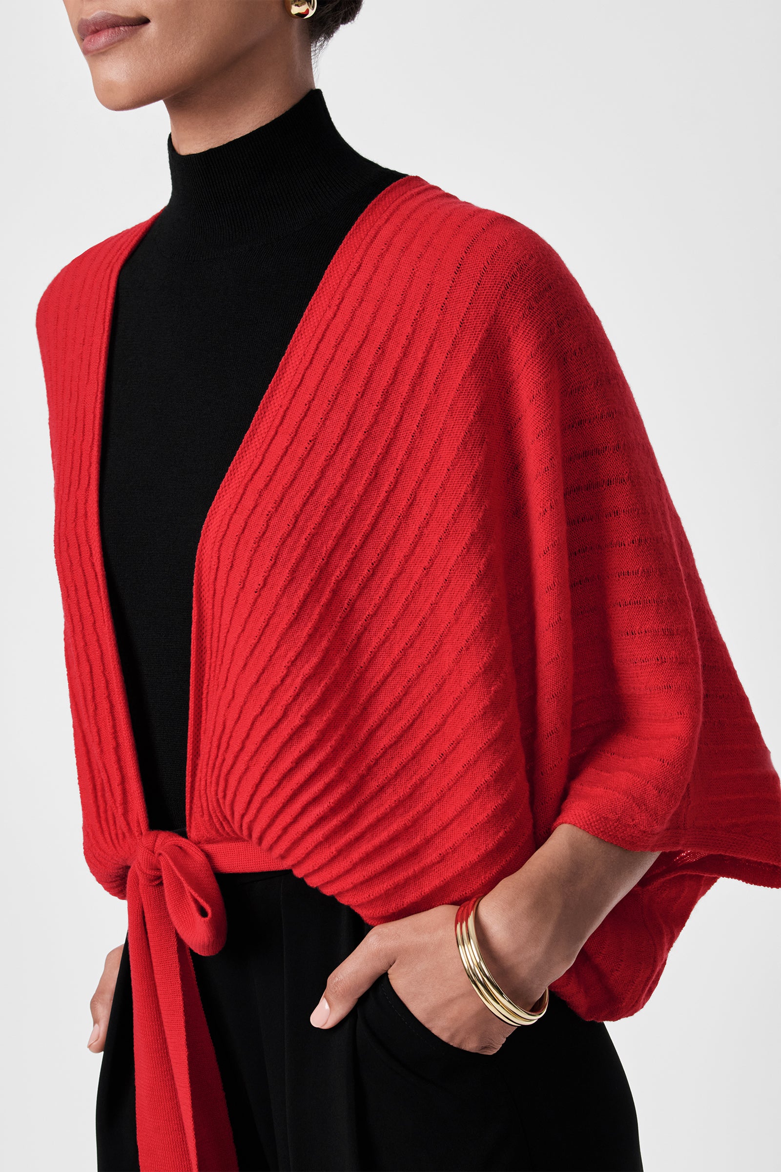 A woman wears the Exceptional Tie Front Sweater in red, tied at the waist over a black turtleneck top and black pants, styled with gold hoop earrings and gold bangles. The backdrop is plain white.