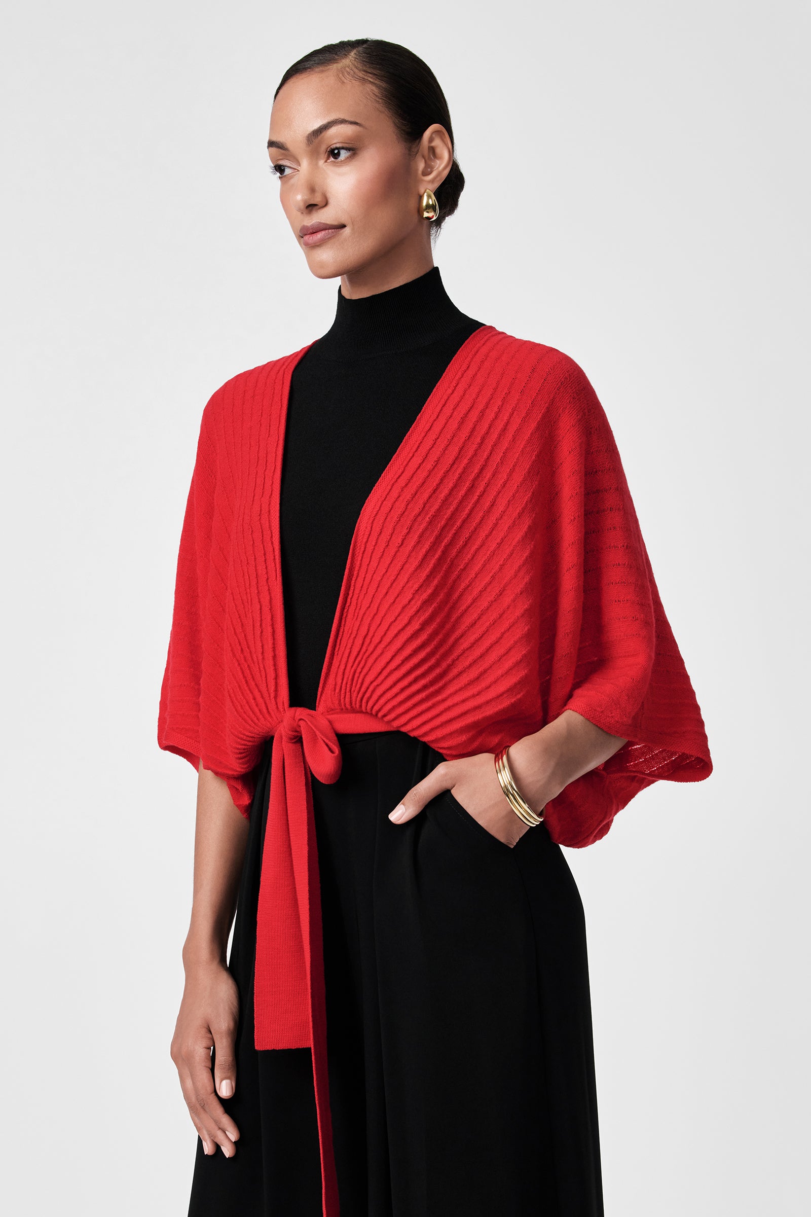 A woman wears the Exceptional Tie Front Sweater in red over a black dress, styled at the waist, with gold hoop earrings and a bracelet, standing with one hand in her pocket against a light gray background.