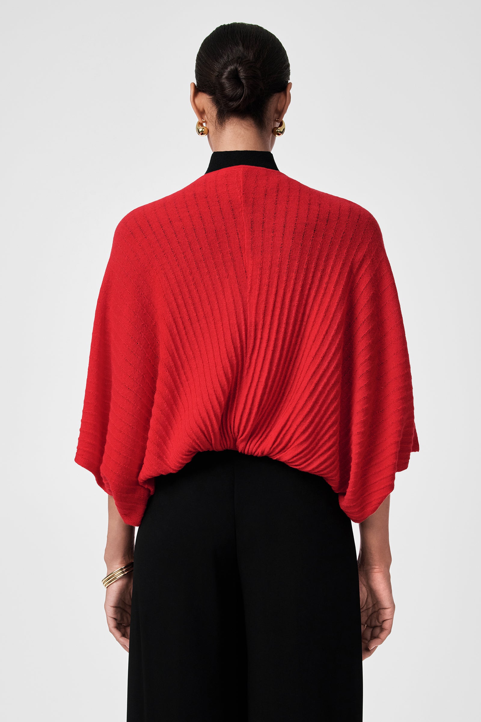 A person stands with their back to the camera, wearing the Exceptional Tie Front Sweater in textured red with wide sleeves, paired with black high-waisted pants. Their hair is in a neat bun, accessorized with gold earrings and bracelets.