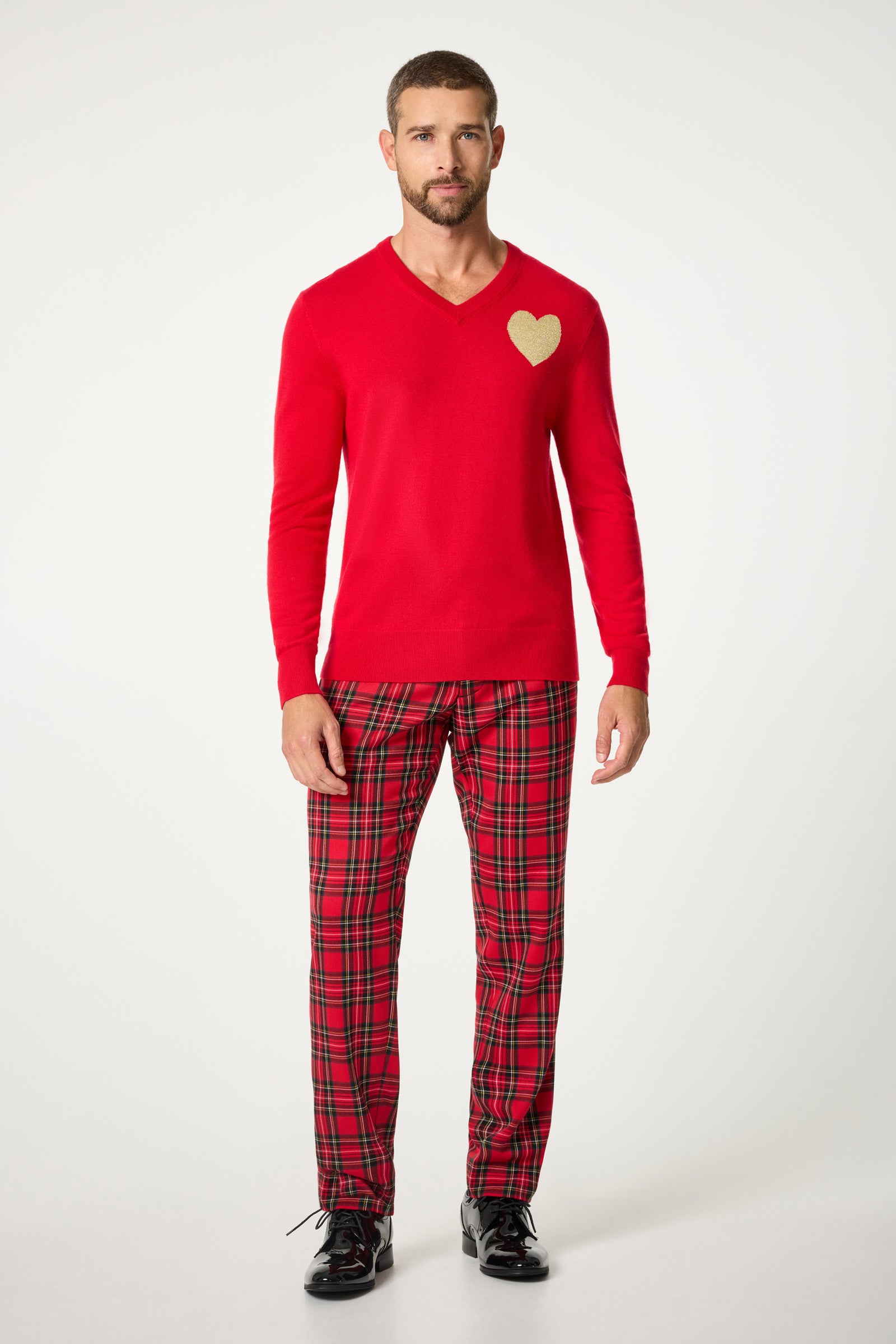 A man with short hair and a beard stands against a plain background wearing the Lover V-Neck Sweater, featuring a gold heart on the chest, red plaid pajama pants, and black shoes.
