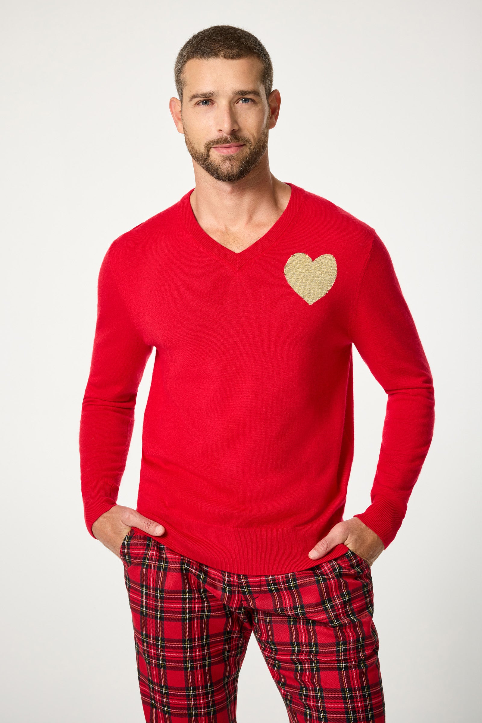 A man with short hair and a beard wears the Lover V-Neck Sweater, a bright red top featuring a gold heart on the chest, paired with red plaid pants. He stands with his hands in his pockets against a simple light background.
