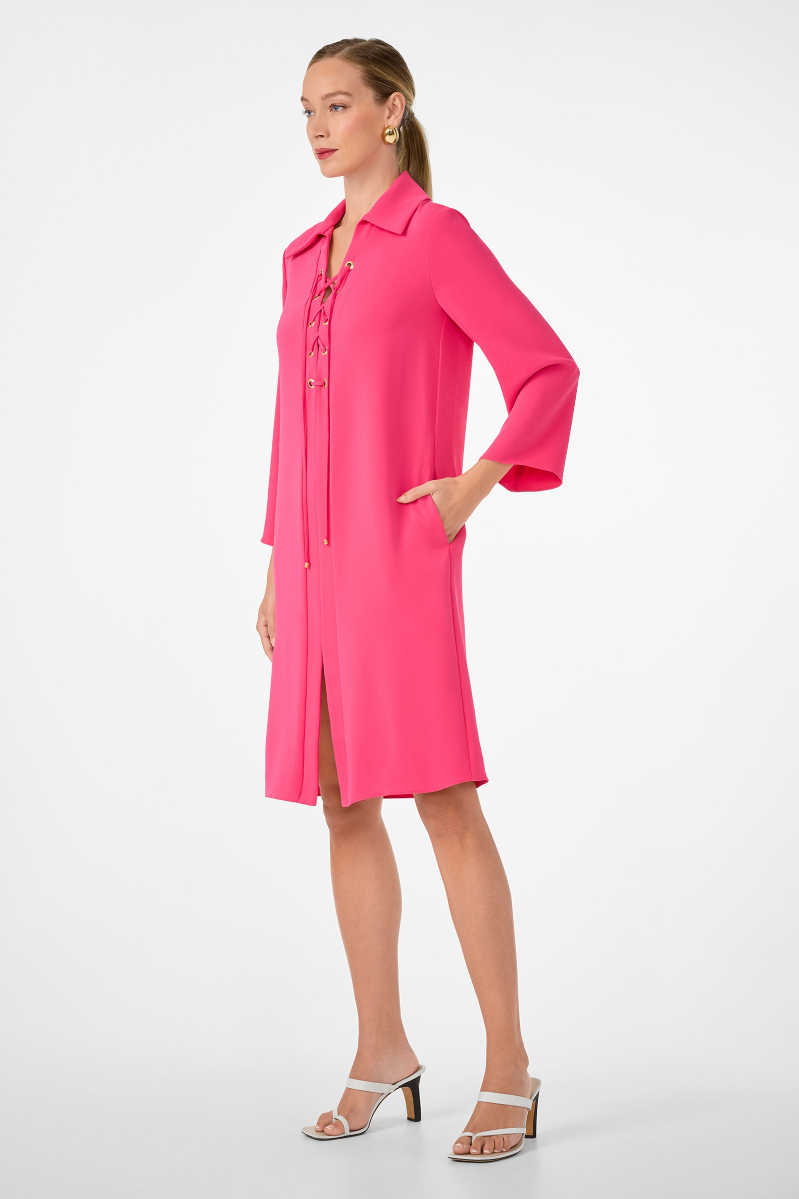 A woman with her hair tied back stands against a plain background, wearing the Janeiro Dress—a bright pink, knee-length dress with a lace-up front and long sleeves—paired with white heeled sandals and one hand in her pocket.