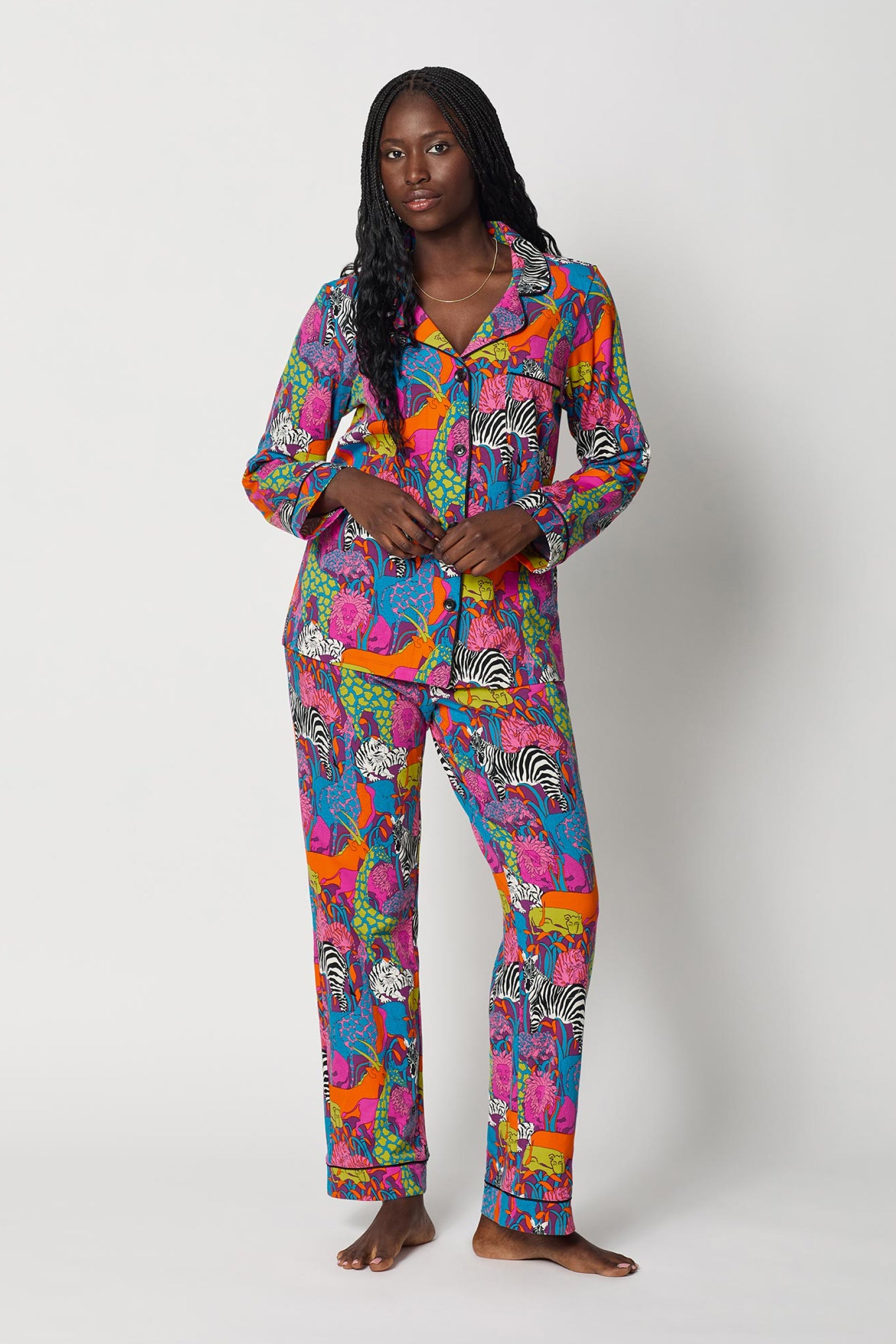 A woman stands barefoot against a plain white background, wearing the Classic PJ Set - Jungle. The pajamas feature bold animal prints like zebras and deer in vibrant shades of pink, blue, orange, and green.