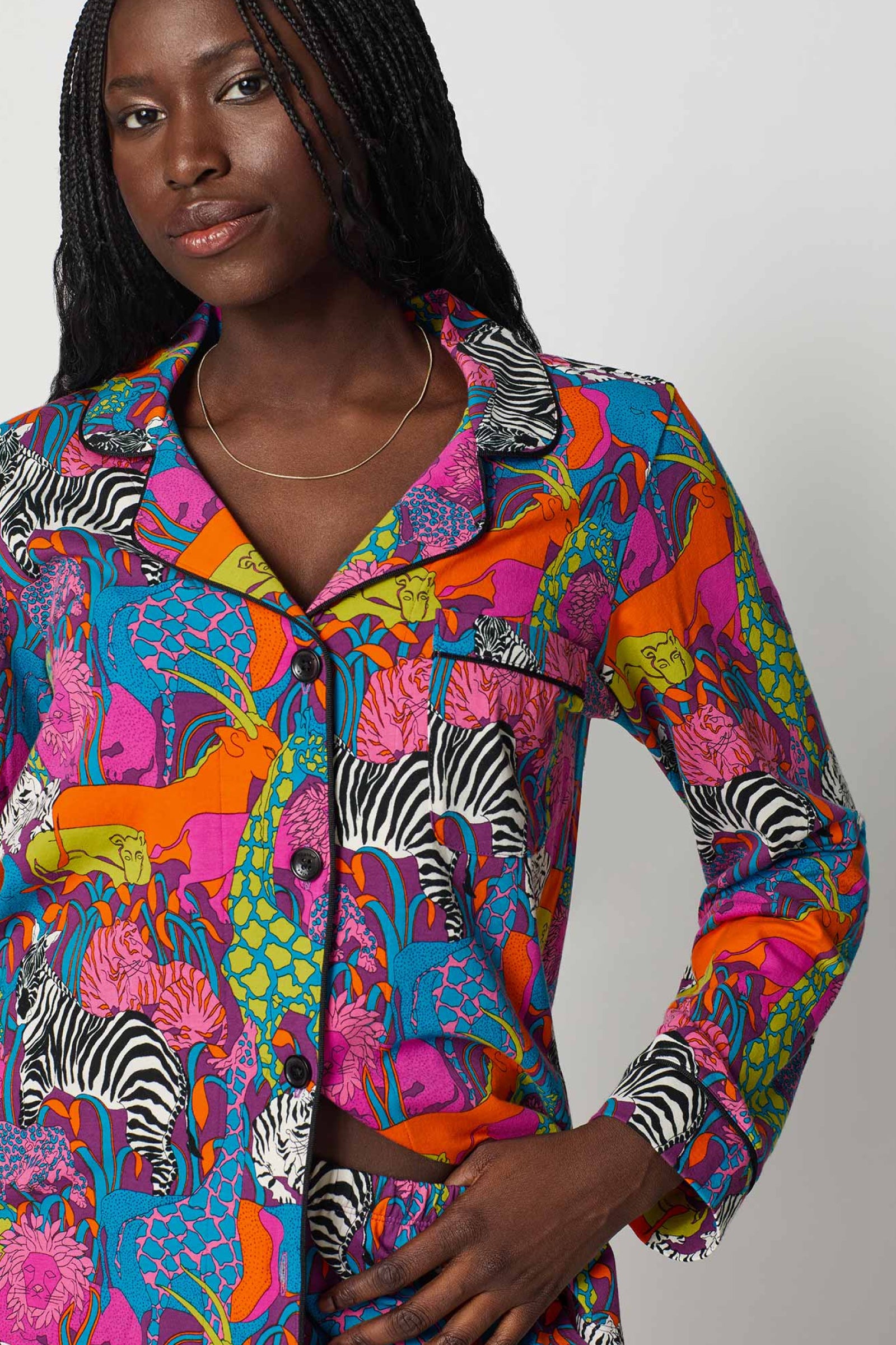 A woman with long braided hair models the Classic PJ Set - Jungle, a colorful button-up featuring vibrant prints of zebras, giraffes, and lions in blue, orange, pink, and purple. She faces forward with one hand on her hip.