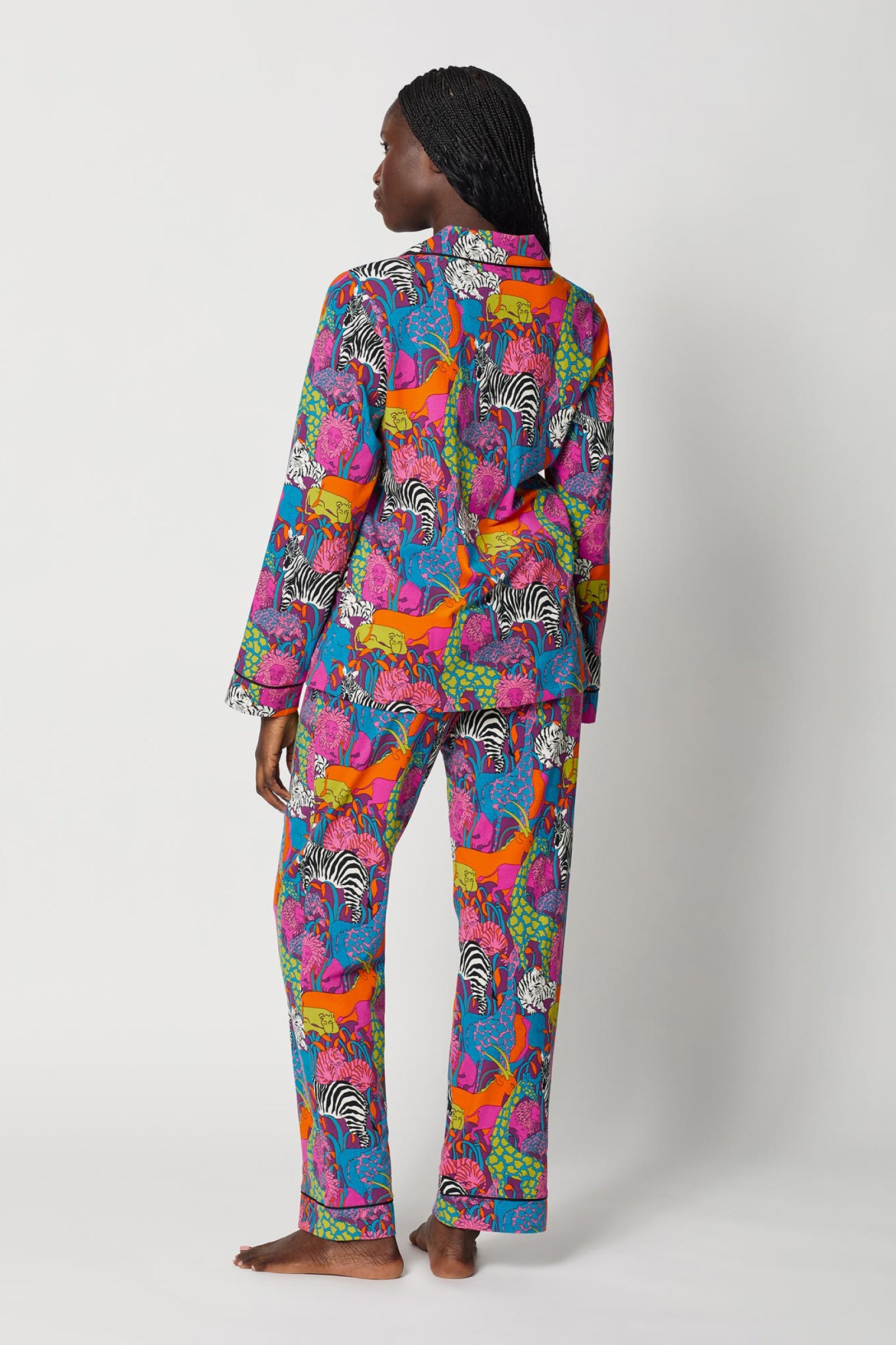 A person stands barefoot and faces away, wearing the Classic PJ Set - Jungle, which features a vibrant animal print with zebras, giraffes, and foliage on a white background.