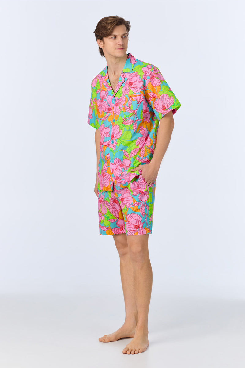 A man stands barefoot wearing the Mens Boxer PJ Set - Linear Floral Poplin, featuring a colorful short-sleeved shirt and matching shorts with a bright pink, green, and blue floral pattern against a plain light background.
