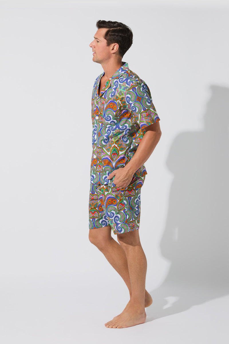 A man stands barefoot against a plain background, smiling and turned slightly to the side, wearing the Mens Boxer PJ Set - Paisley: a vibrant, colorful matching short-sleeve shirt and shorts with a bold paisley pattern.