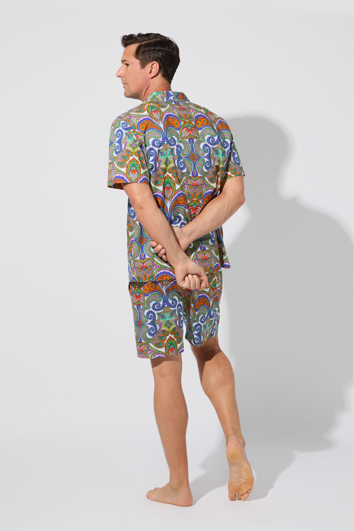 A man stands barefoot on one foot, facing away, wearing the Mens Boxer PJ Set - Paisley with a colorful pattern. He has his arms behind his back in a studio with a plain white background.