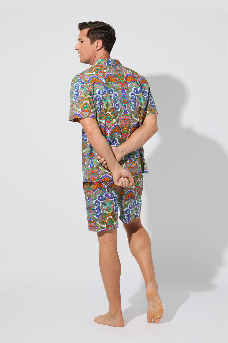 A man stands barefoot on one foot, facing away, wearing the Mens Boxer PJ Set - Paisley with a colorful pattern. He has his arms behind his back in a studio with a plain white background.