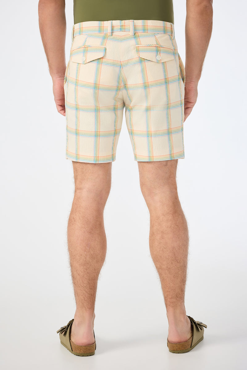 A person stands facing away, wearing the Travis Short - Pride Plaid (beige with blue and orange stripes), a green tucked-in shirt, and brown moccasins against a plain white background.