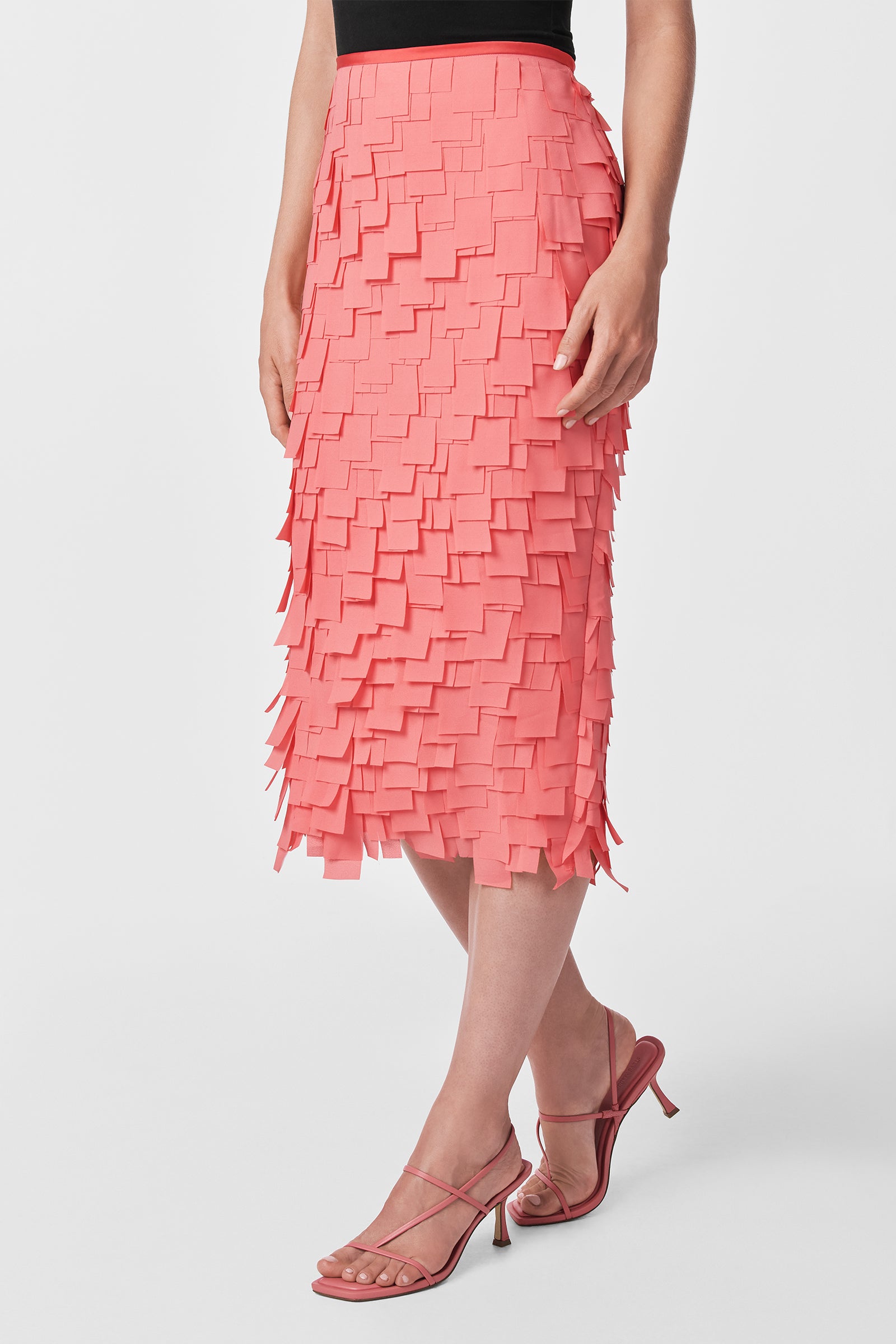 A woman wears the Dita Midi Pencil Skirt - Flutter Chiffon in coral pink with layered square panels, paired with a black top and strappy pink heeled sandals, standing against a plain white background.