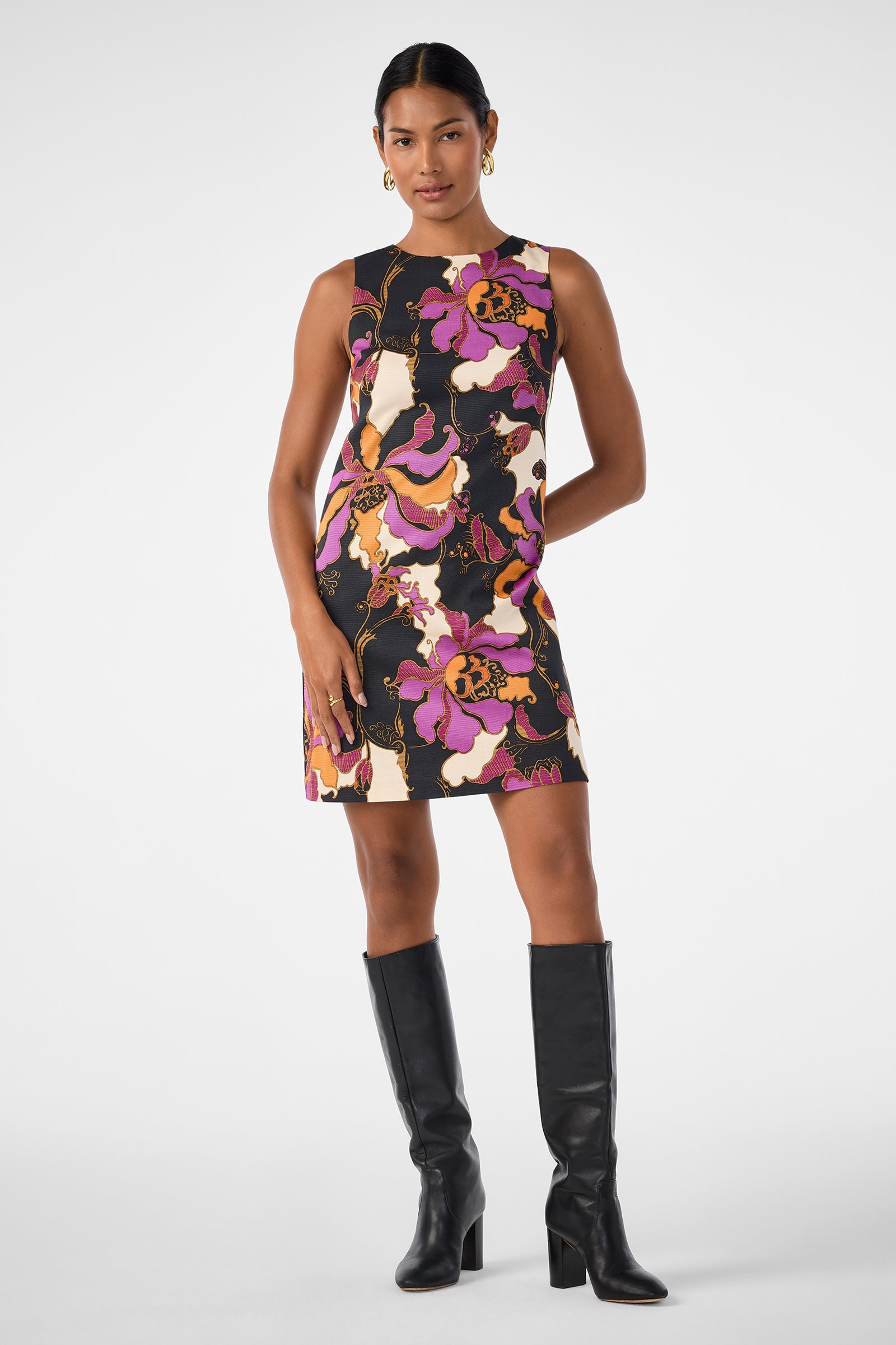 A woman wears the Addy Dress, a sleeveless, knee-length piece with a bold purple, orange, yellow, and black floral pattern. She pairs it with black knee-high boots and gold hoop earrings, posing confidently against a plain background.