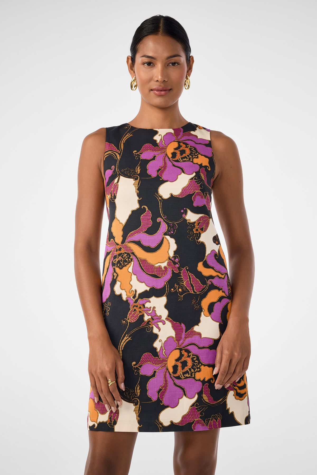 A woman faces forward wearing the Addy Dress, a sleeveless, knee-length design featuring a bold purple, orange, black, and white floral pattern. She has her dark hair pulled back and accessorizes with gold hoop earrings.