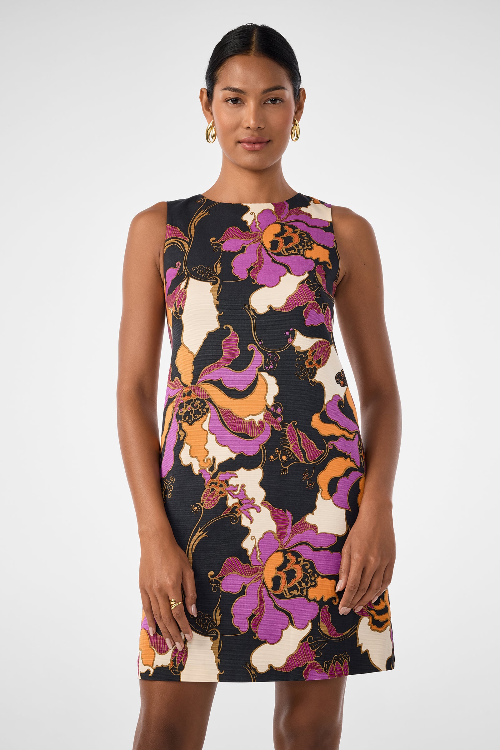 A woman faces forward wearing the Addy Dress, a sleeveless, knee-length design featuring a bold purple, orange, black, and white floral pattern. She has her dark hair pulled back and accessorizes with gold hoop earrings.