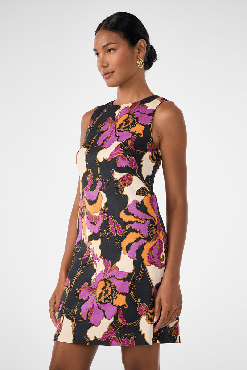 A woman models the Addy Dress—a sleeveless, fitted style with a bold floral print in purple, orange, black, and cream—her hair pulled back as she poses against a plain light background.
