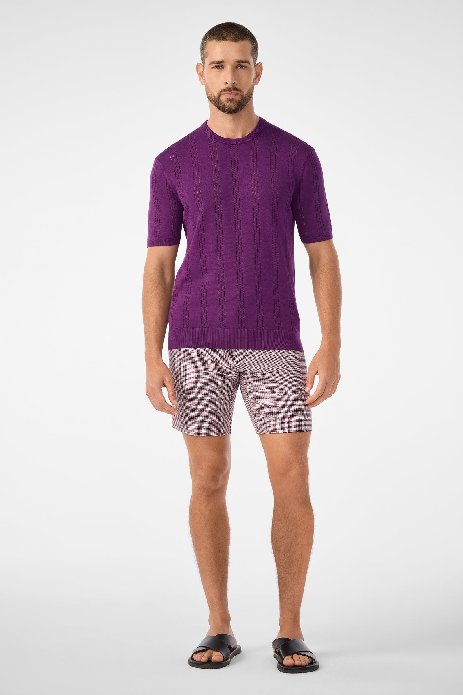 A man stands facing forward, wearing the Lawrence Short, a short-sleeved purple knit sweater, and black sandals against a plain white background.