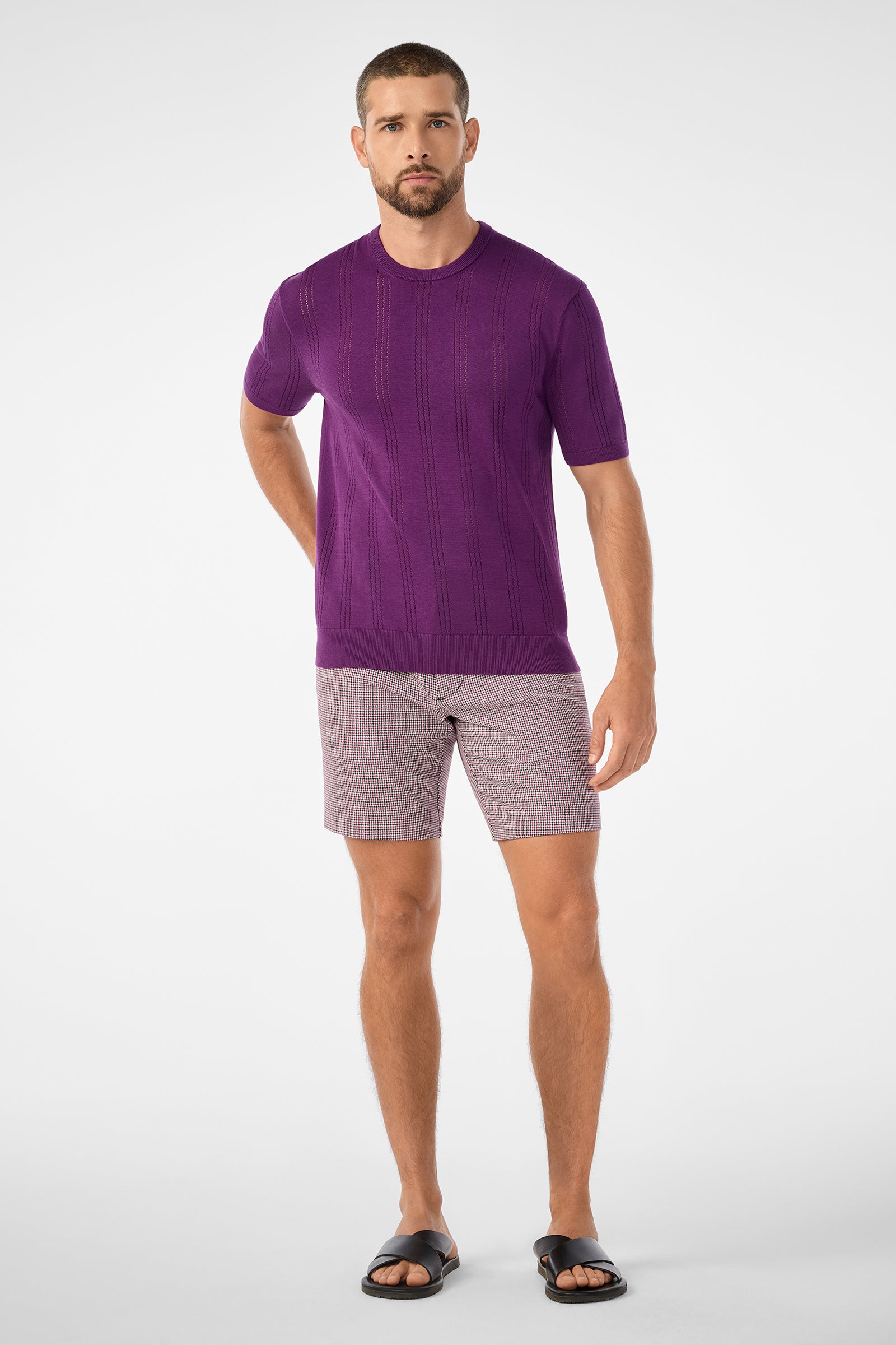 A man with short hair, a beard, and a neutral expression stands against a plain background wearing the Carlo Short-Sleeve Crewneck in purple, plaid shorts, and black slide sandals.
