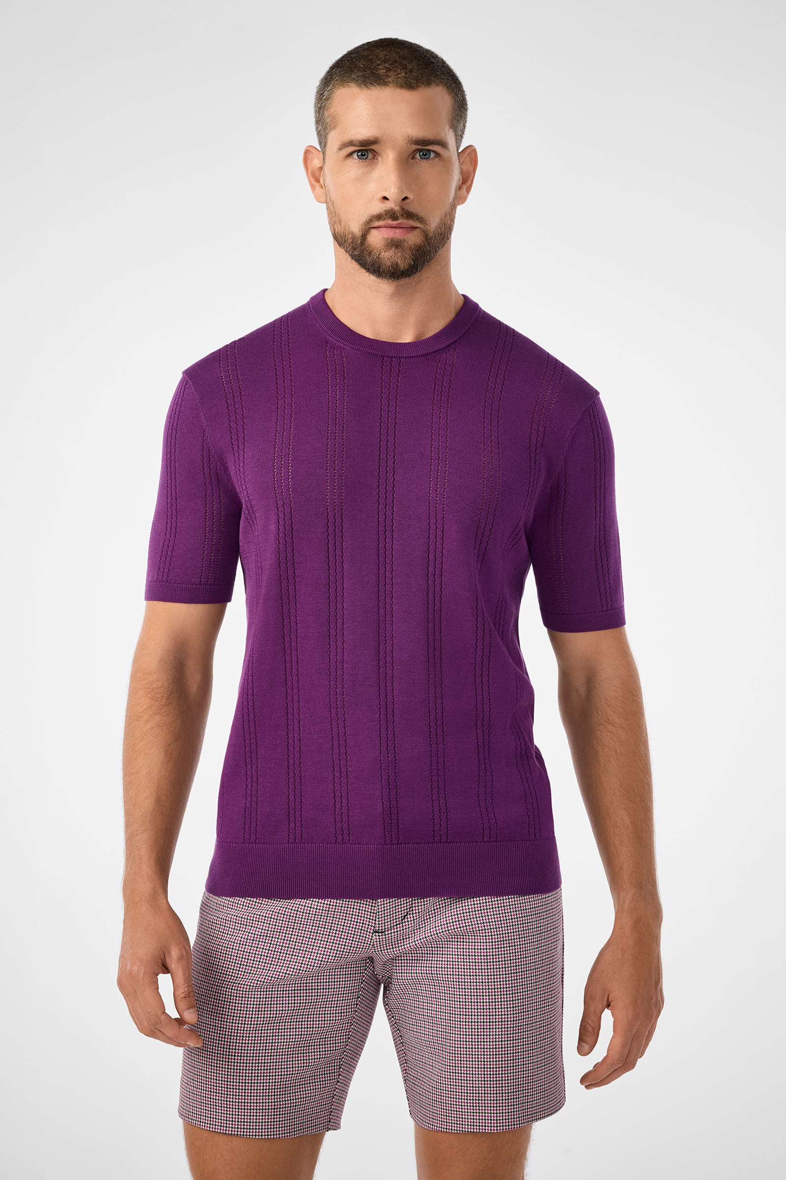 A man with short brown hair and a beard wears the fitted purple Carlo Short-Sleeve Crewneck sweater and patterned shorts, standing against a plain white background.