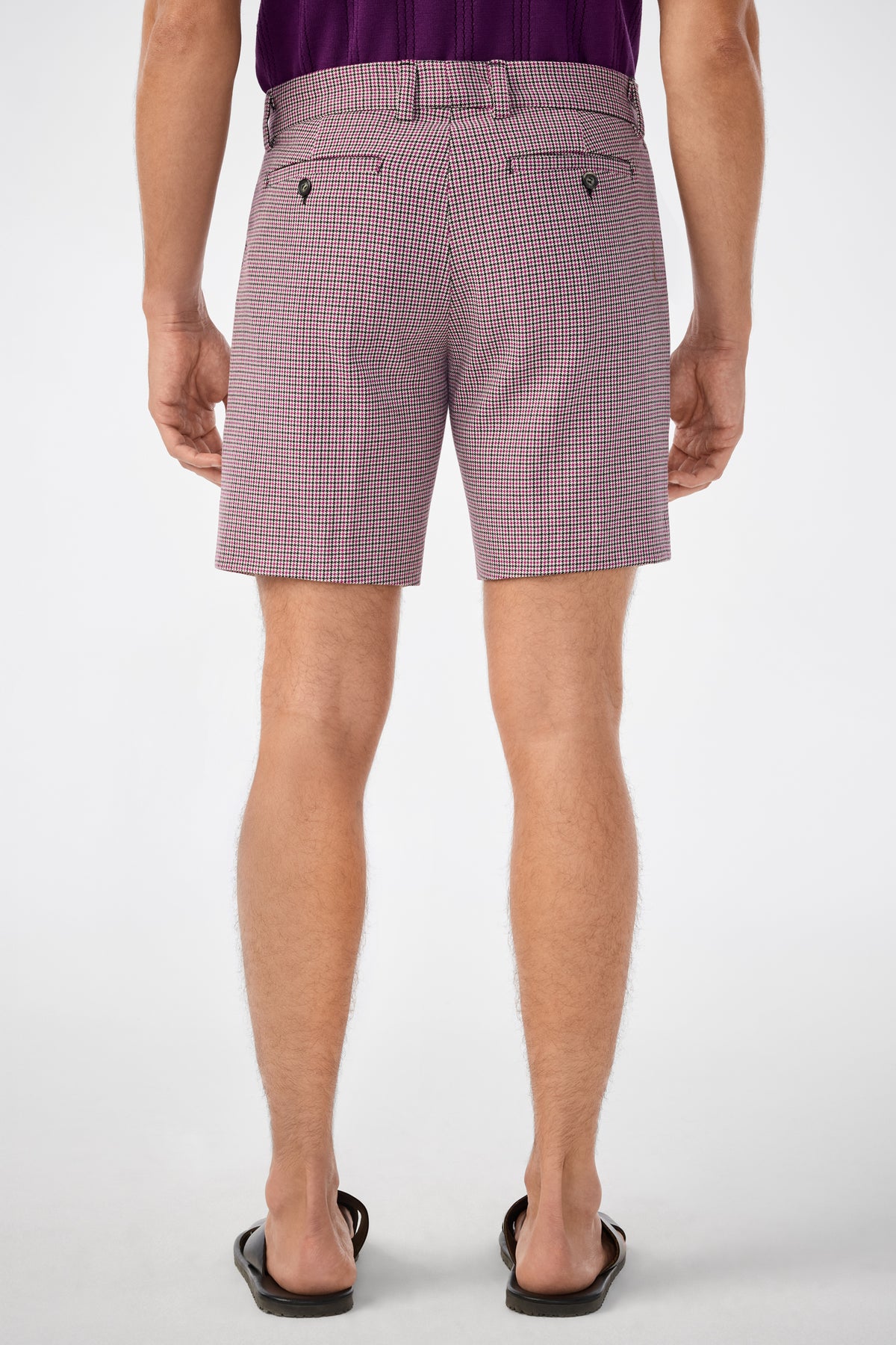 Someone stands facing away in a light background, wearing checkered Lawrence Shorts with buttoned back pockets, a purple shirt, and black flip-flops. The focus is on the back view of the shorts.