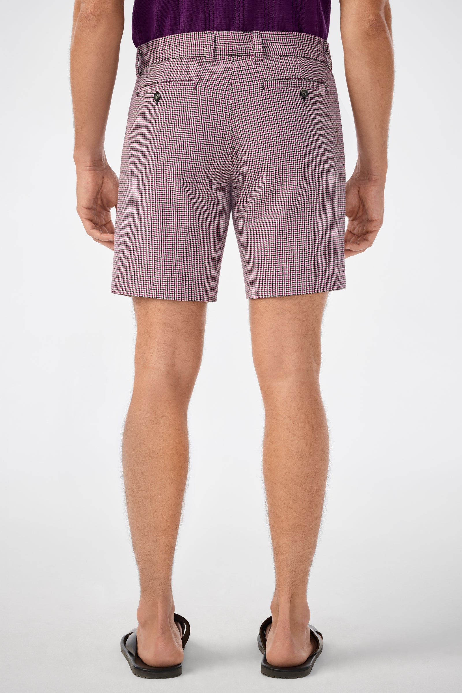 Someone stands facing away in a light background, wearing checkered Lawrence Shorts with buttoned back pockets, a purple shirt, and black flip-flops. The focus is on the back view of the shorts.