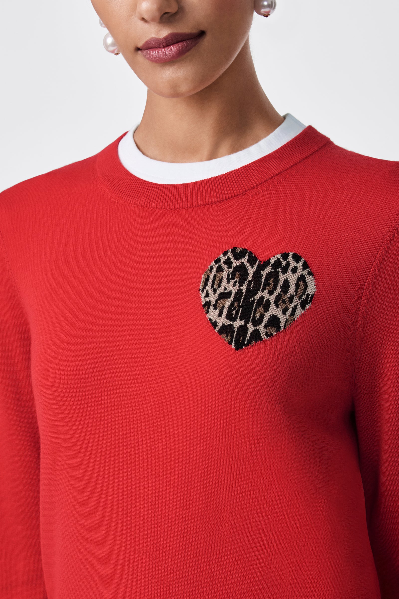 A woman wears the Jaguar Heart Crewneck Sweater, featuring a red color, white neckline, and a heart-shaped leopard print patch on the left chest. Only her lower face and upper torso are shown.